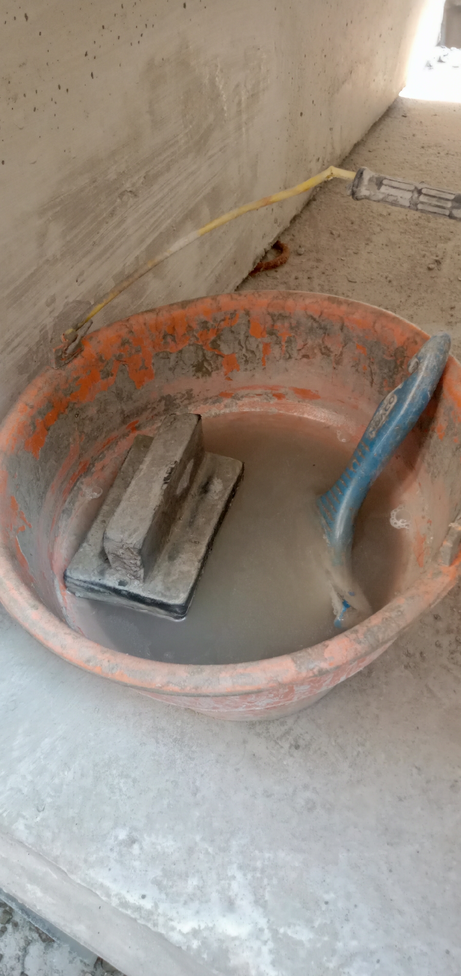 The image depicts a construction scene. An orange bucket contains a gray liquid, likely water or a solution used for cleaning or mixing materials. Inside the bucket, there is a gray sponge or grout float and a blue plastic trowel. The bucket is positioned against a gray concrete wall. A yellow hose runs along the wall, extending into the frame from the upper right. The scene appears to be indoors or in a sheltered area, possibly a construction site. The location is unknown.