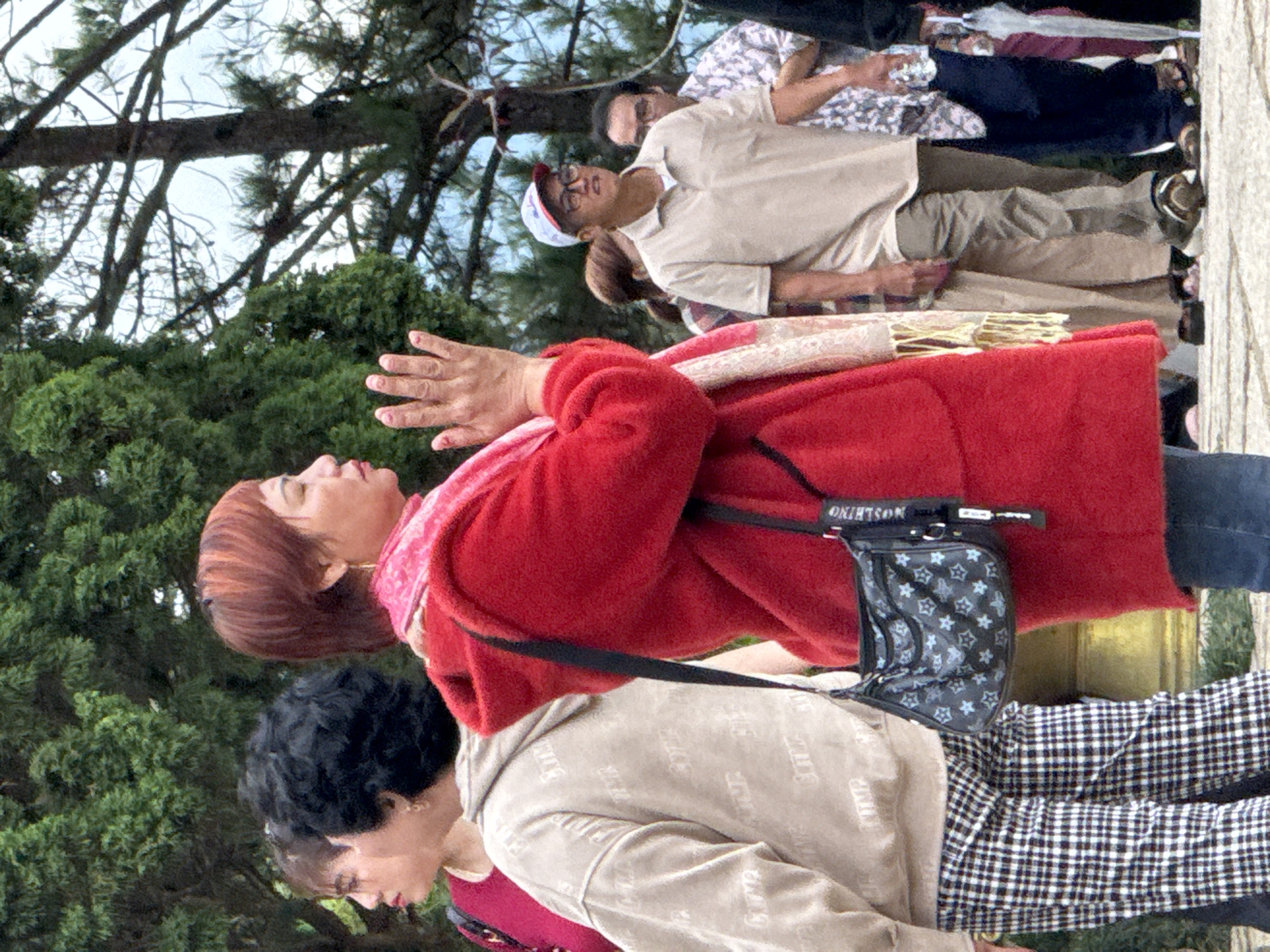 The image depicts a group of people outdoors, possibly at a tourist attraction or religious site. The focal point is a woman in the foreground wearing a red coat and a pink scarf, with her hands clasped in front of her. She has short, reddish-brown hair. To her left, another woman with dark, curly hair is partially visible, wearing a light-colored hooded jacket. In the background, several other individuals are seen, including a man wearing a light brown polo shirt and a baseball cap. The environment appears to be a park or garden with abundant greenery and trees. The architecture in the background, a large golden structure, is characteristic of some temples or pagodas found in Vietnam, and the presence of lush vegetation suggests a location like the Tuyen Lam Lake area or a similar scenic spot in Da Lat, Vietnam.