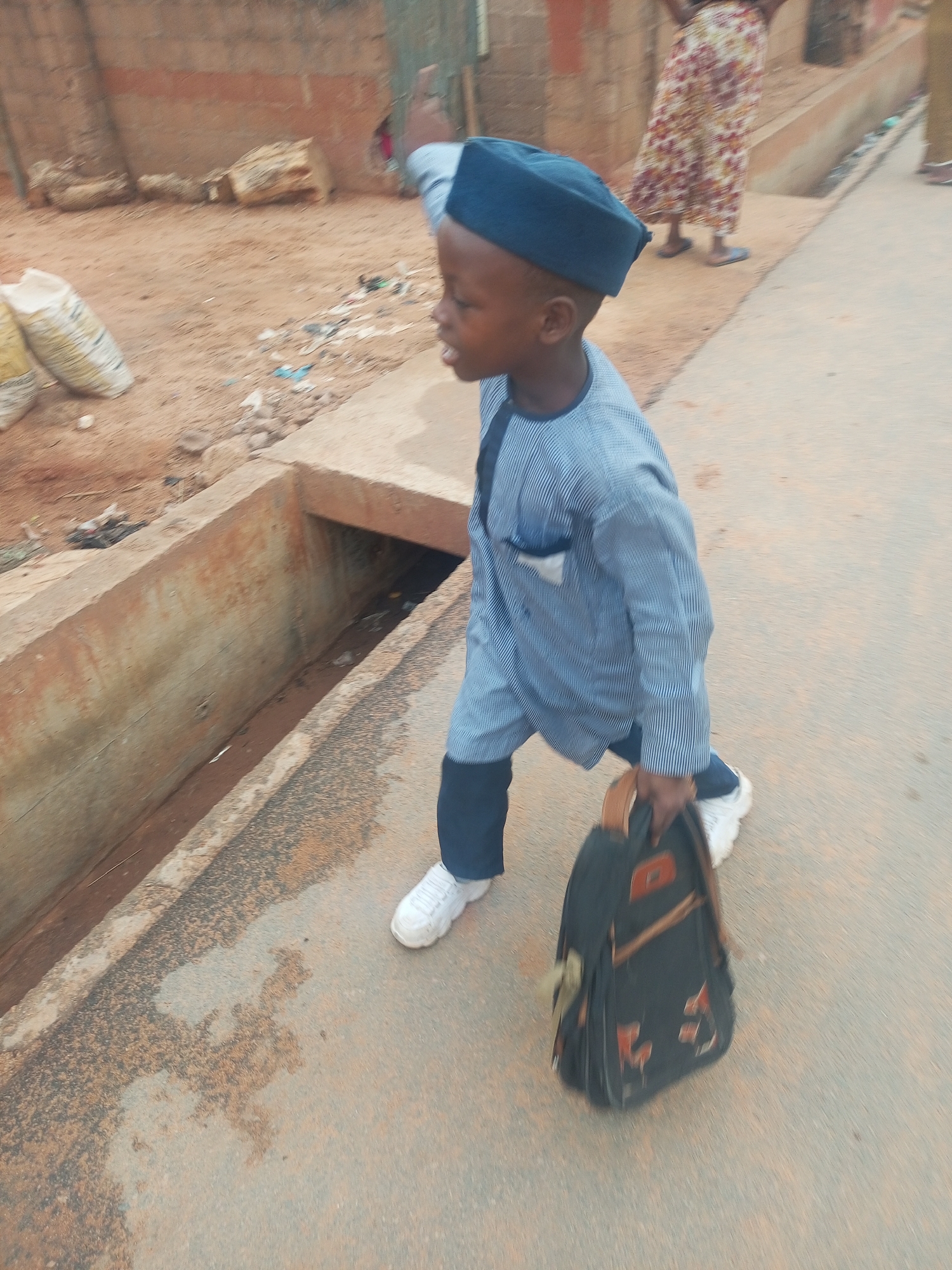 A young boy dressed in a blue striped suit and matching cap walks on a paved surface, carrying a dark backpack with orange accents. He is wearing white sneakers. The environment appears to be a street in Tempure, Nigeria, with a concrete drainage ditch to his left. In the background, a woman in a patterned dress is partially visible. There are bags and debris on the unpaved ground behind the ditch.