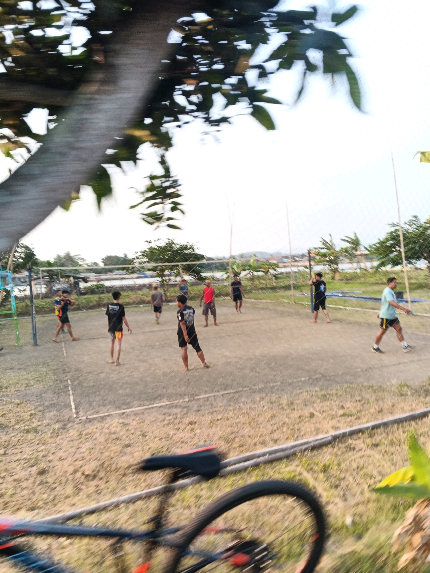 The image depicts an outdoor volleyball game in progress on a dirt court, possibly near a body of water, given the presence of what appears to be houses and vegetation in the background consistent with the Cilacap region of Indonesia. Several young men are actively playing volleyball. One individual in a light blue shirt and dark shorts is walking towards the left of the frame. Another person wearing a black shirt with a graphic design and dark shorts is in motion on the right side of the court. A person in a black t-shirt with colorful shorts stands in the middle of the court. To the left, two individuals are visible on the court; one is wearing a black and yellow shirt with shorts, and another in a black shirt and shorts. Further back, a person in a red shirt and shorts is positioned on the right side of the court, while another individual in a grey shirt and dark shorts is visible in the background. The court is enclosed by a net, and several poles support it. In the foreground, a bicycle wheel and part of its frame are blurred due to motion, suggesting it was either moving or the photograph was taken with a moving camera. The environment shows dry grass and trees, indicative of a tropical climate.