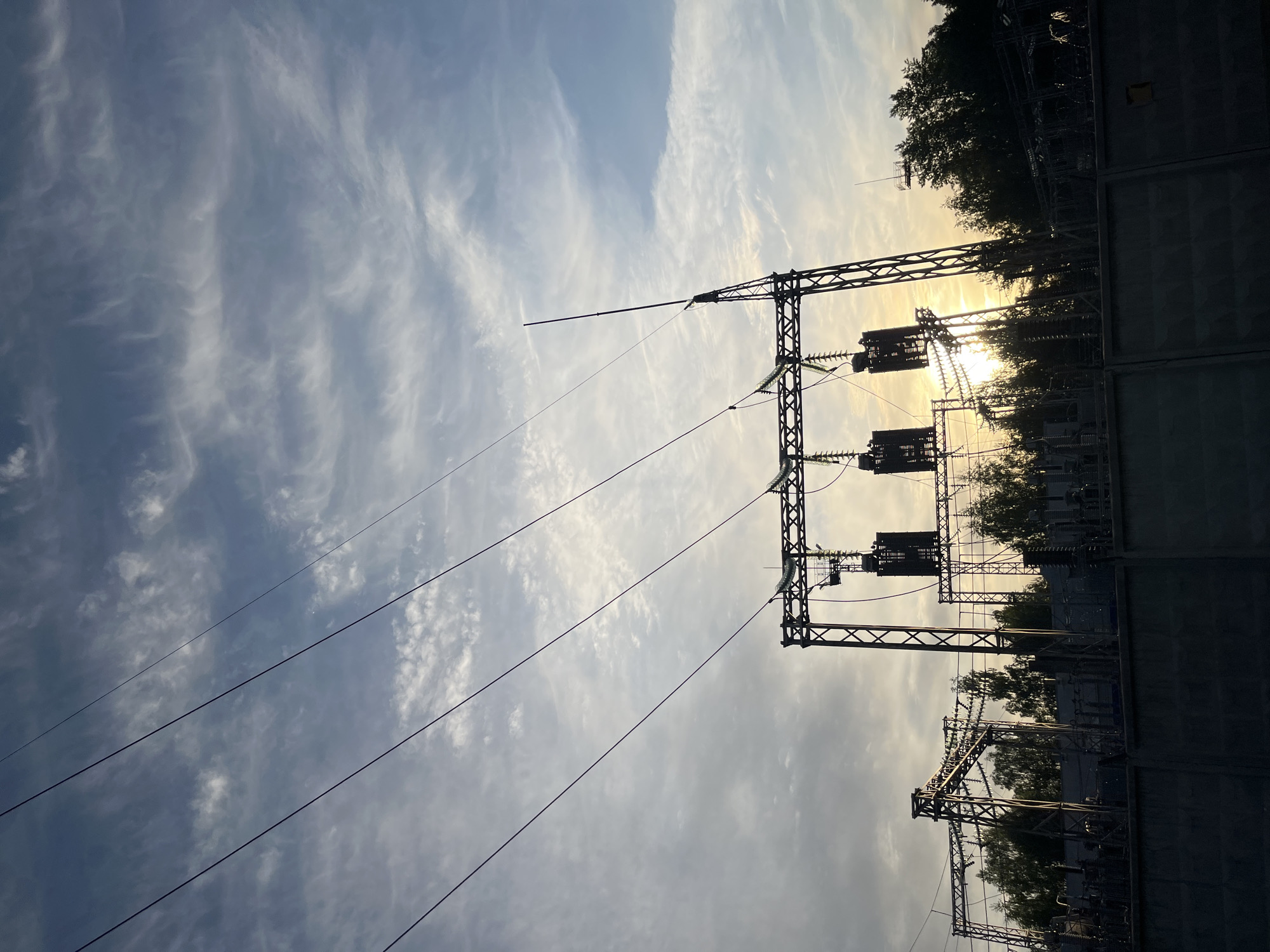 This image shows a power substation in Chaikovsky, Russia, photographed during the late afternoon or early evening. The power lines and electrical towers dominate the scene, silhouetted against the sun and a sky filled with cirrus clouds. The sun is partially obscured by trees, casting a bright glare. The scene appears to be on the outskirts of the city, as suggested by the combination of industrial infrastructure and natural elements. The absence of people indicates the area is likely secured. The image is rotated counter-clockwise by approximately 90 degrees.