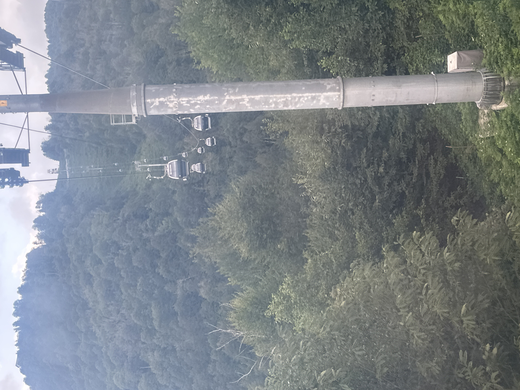 The image depicts a view from a ski lift or gondola in Esto-Sadok, Russia, ascending a densely forested mountainside. Several gondolas are visible on the cable, moving up the slope. A large, grey metal pylon for the cableway dominates the foreground on the right side of the image. The surrounding environment is characterized by thick green trees covering the mountain, with some lighter green foliage in the foreground. The sky above is overcast with a hazy, muted light. A small structure, possibly an electrical box, is visible at the base of the pylon.
