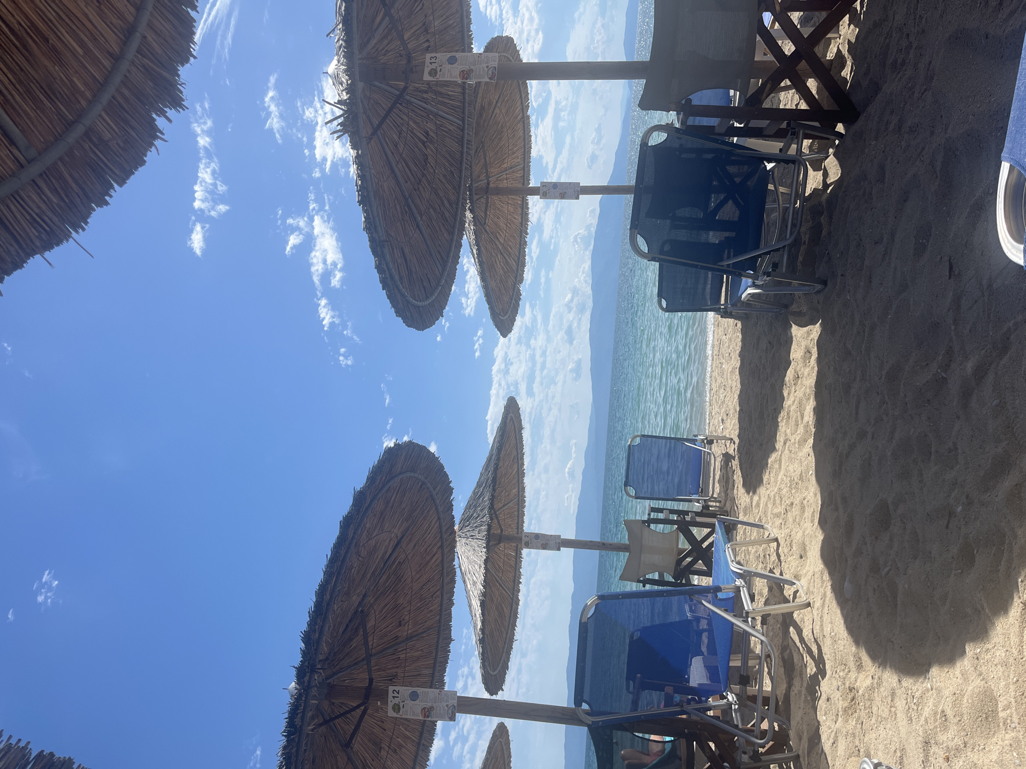 The image shows a beach scene at Ofrynio Beach, Greece, on a clear day with a bright blue sky and scattered clouds. Several thatched umbrellas provide shade over rows of empty lounge chairs on a sandy beach. The turquoise water of the sea is visible behind the chairs, with mountains in the distance. The lounge chairs are made of metal and fabric, with some in blue and others in neutral colors. Signs are attached to the poles of some umbrellas, displaying numbers and what appear to be menus or information. The sand is light brown and shows shadows from the umbrellas and chairs. The overall atmosphere is peaceful and inviting, typical of a beach resort.