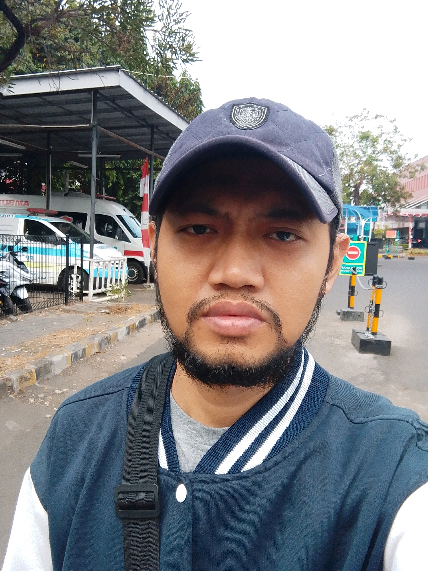 A medium shot shows a man wearing a blue baseball cap with a crest and a blue bomber jacket over a grey t-shirt, with a black strap across his chest. He is standing outside, facing the camera, with a slight smile. Behind him, two white ambulances with red and blue markings are parked under a metal awning. A motorcycle is parked to the left of the ambulances. The background shows a street with a sidewalk, a traffic sign with a red circle and line, and buildings. The scene appears to be in a public area, possibly a hospital or clinic parking lot. The presence of ambulances and the Indonesian flag suggest a location in Indonesia, and the style of the buildings and vehicles is consistent with East Jakarta.