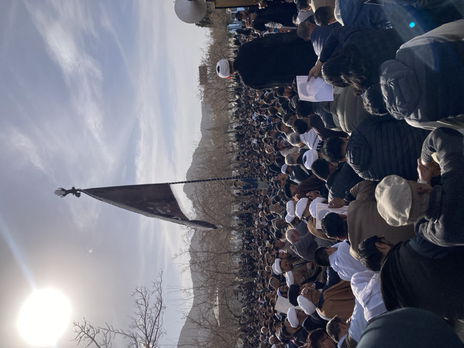 An outdoor scene in Quetta City Tehsil, Pakistan, depicts a large congregation of individuals, predominantly male, gathered in what appears to be an open public area. The sky is bright with direct sunlight and scattered clouds. In the background, barren deciduous trees and distant mountains are visible.
A prominent feature in the center-left of the image is a tall black flag, bearing indistinct white script or symbols, mounted on a pole. The flagpole is topped with a decorative finial shaped like an open hand. To the far right, a large white horn-shaped loudspeaker is visible, mounted on an elevated structure.
The crowd consists of hundreds of individuals, most of whom are seated on the ground, facing generally towards the front (viewer's perspective is from the rear of the crowd). Many attendees wear traditional attire, including various styles of turbans (white and light brown) and skullcaps. In the mid-ground, a man wearing a black robe and a white turban stands, facing away from the camera, seemingly addressing or observing the crowd.
Several individuals within the crowd are holding up printed white sheets of paper, each displaying a portrait of an elderly man with a beard. The overall atmosphere suggests an organized gathering, possibly a religious or commemorative event.