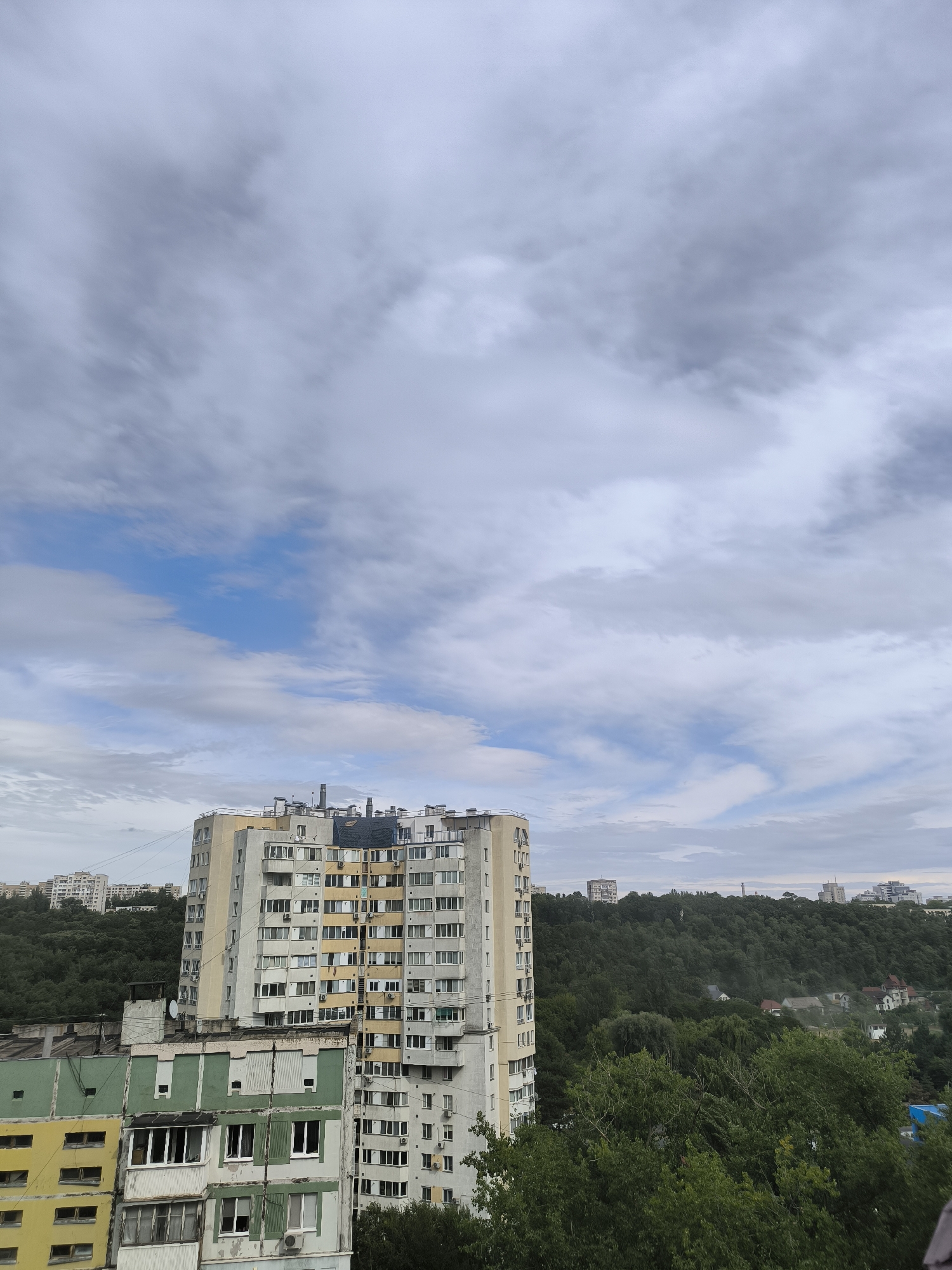 The image captures an overcast sky with scattered clouds above a cityscape. Dominating the center is a multi-story residential building, likely an apartment block, characterized by its numerous windows and balconies. Adjacent to it, to the left, is another building with distinct green and yellow sections. In the background, a densely wooded hillside separates the foreground buildings from a more distant cluster of structures, suggesting a city or town built on undulating terrain. The overall impression is one of a residential area with a significant amount of greenery. Given the architectural style and the presence of similar buildings in other available imagery, this scene is located in the Râșcani sector of Chișinău, Moldova, specifically near the intersection of Kiev street and Moscow Avenue, an area known for its Soviet-era apartment blocks and surrounding green spaces. No people are visible in the image, and the scene depicts a static urban environment.