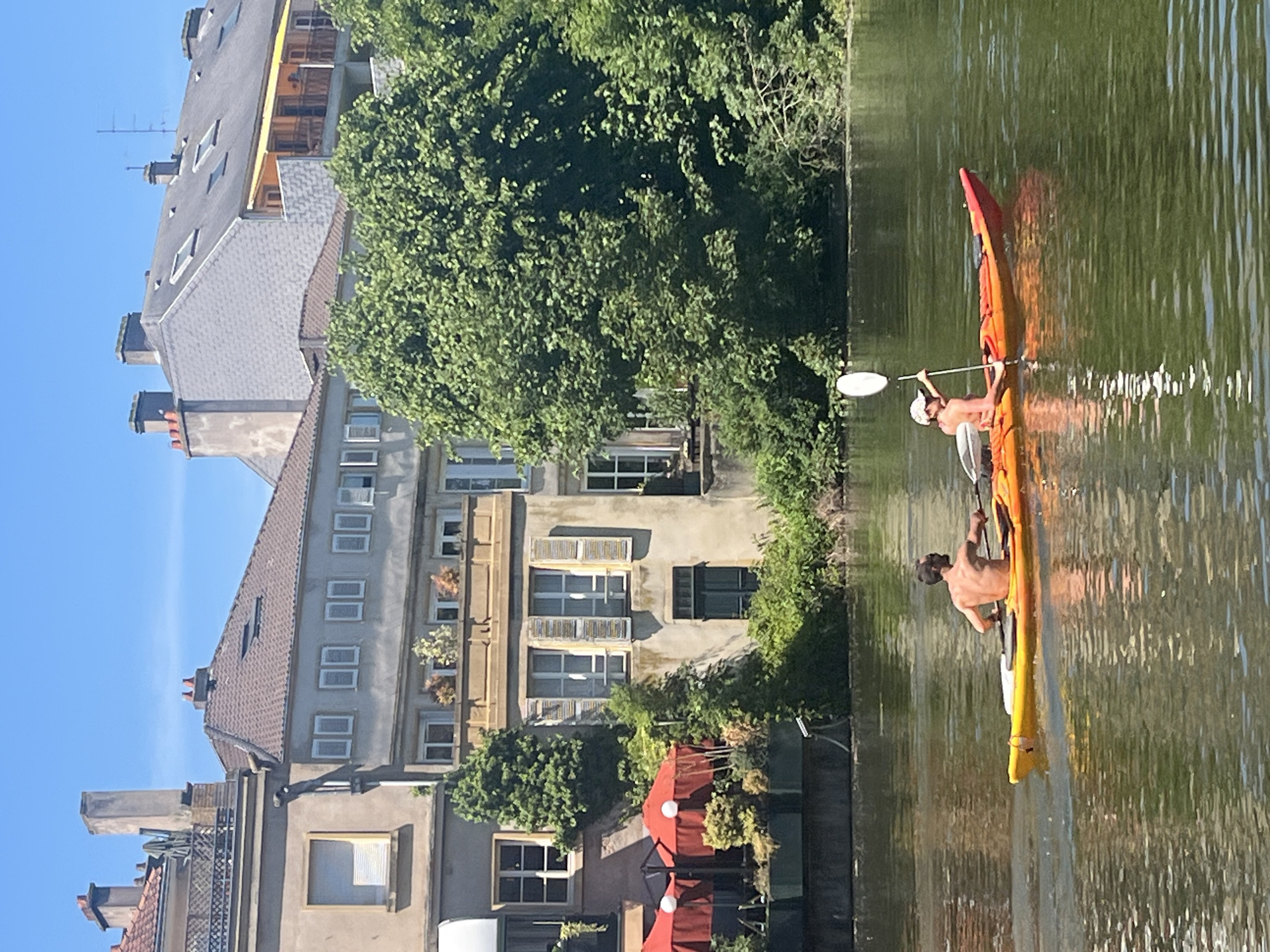 Two shirtless men are kayaking on a canal in Metz, France. One man is in the front of the orange and yellow kayak, paddling with a white paddle. The other man is in the back, also paddling. The canal is surrounded by buildings, trees, and shrubbery. The buildings are old and have many windows. Some of the windows have shutters. There is a red awning visible in front of one of the buildings. The sky is blue and clear. The water is calm.