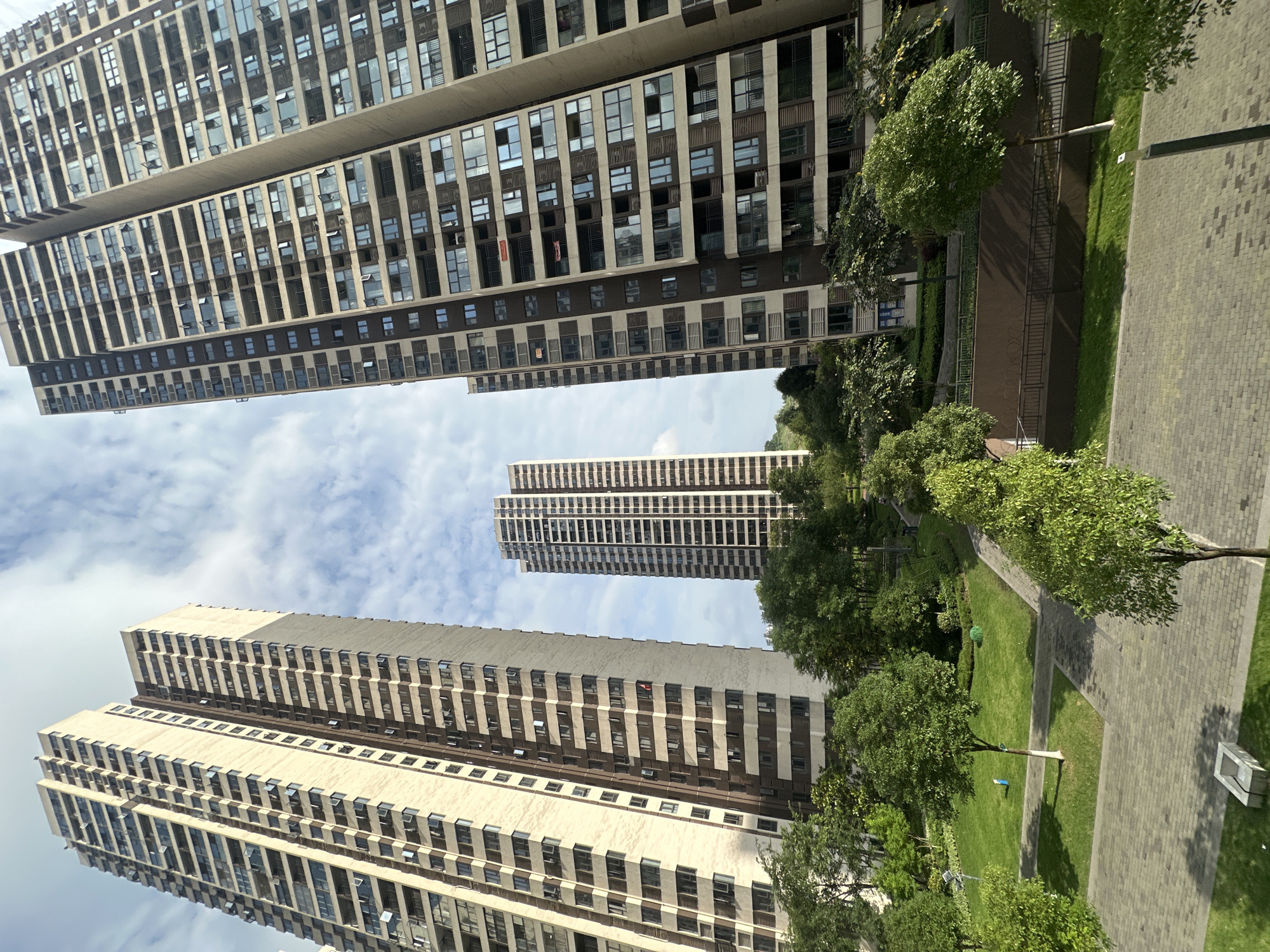 The image depicts a view in Chongqing High-tech Zone, China, with several high-rise apartment buildings framing the sky. The buildings are constructed of light-colored materials with multiple rows of windows, creating a grid-like pattern on their facades. Between the buildings, there is a glimpse of a partly cloudy sky. Below, a park area is visible, with green lawns, trees, and stone paved walkways. The perspective is tilted upward, emphasizing the height of the buildings. There are no people visible in the frame.