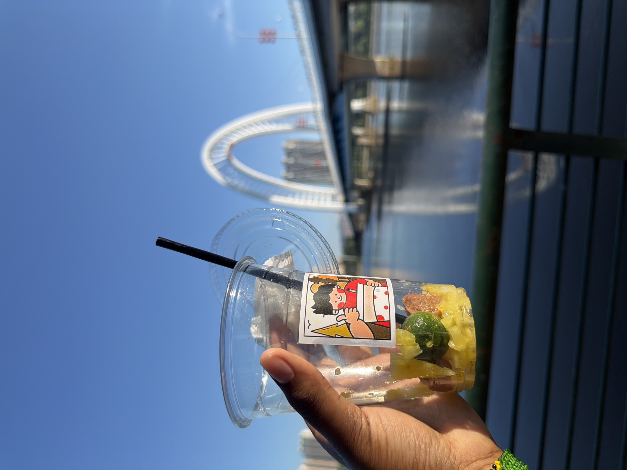 In Linghe, China, a person's hand holds a clear plastic cup containing a drink with chunks of fruit and what appear to be small pieces of meat, along with a green garnish. A black straw extends from the cup, and the lid rests to the side. A decorative sticker on the cup depicts a cartoon character giving a thumbs-up. The background shows a large, arched white bridge spanning a wide body of water under a clear blue sky. Buildings are visible in the distance behind the bridge. The scene is captured outdoors, with a railing in the foreground.