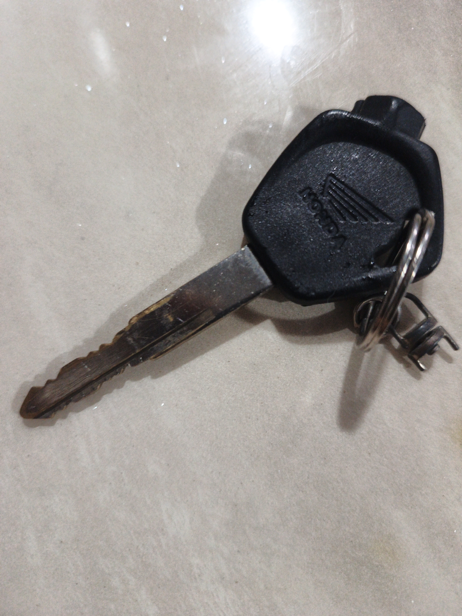 A Honda key is lying on a textured, light-colored surface. The key has a black plastic head with the Honda logo embossed on it. The metal blade of the key is visible and appears worn. A metal keychain is attached to the head of the key, with two rings and what looks like a small metal tag. The surface beneath the key has a slight sheen and appears to be a countertop or floor. The lighting suggests an indoor setting. There are no people or animals visible in the image. The image is taken from a slightly elevated angle, looking down at the key.