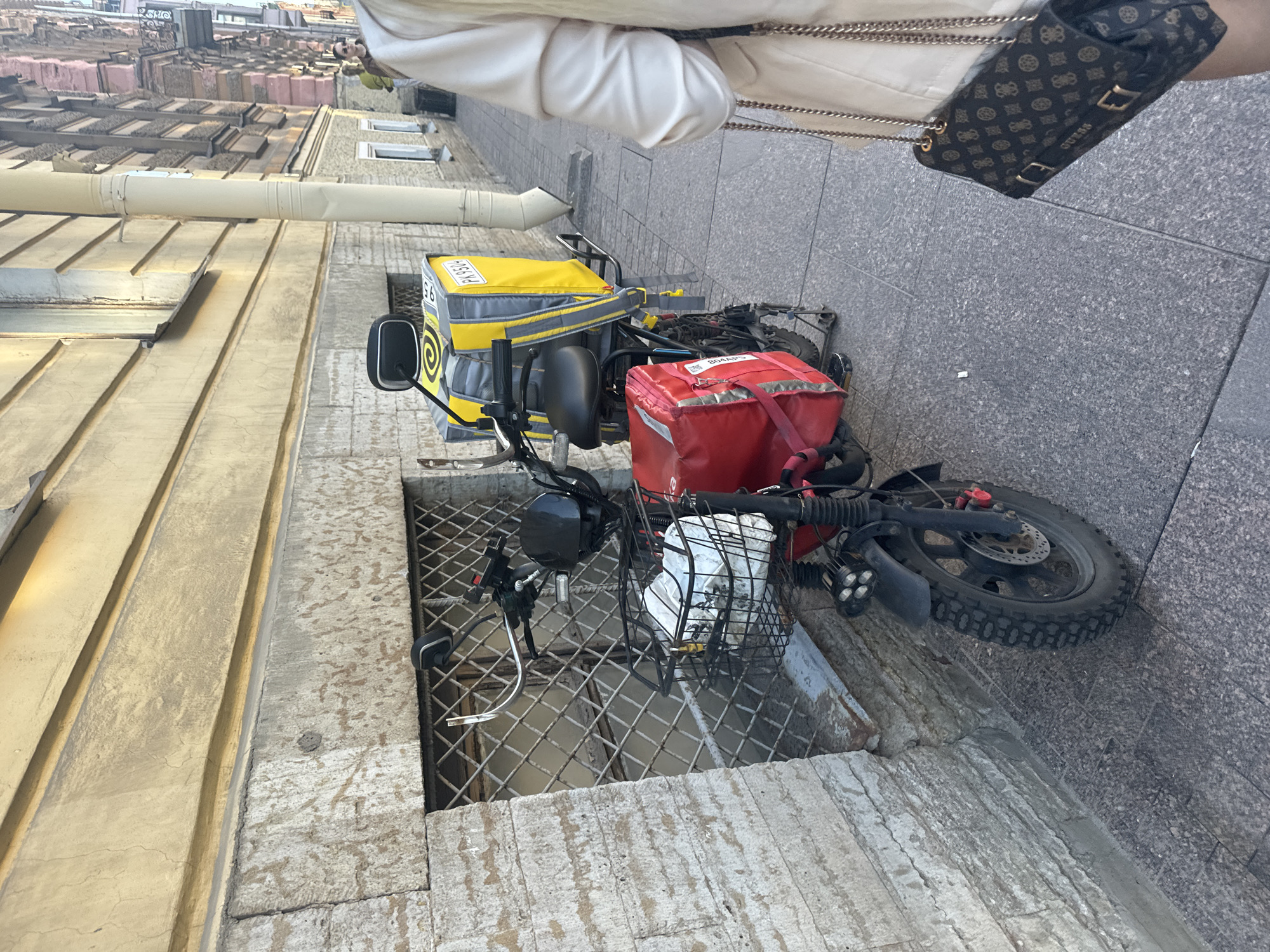 The image depicts a street scene in Saint Petersburg, Russia, focusing on a delivery scooter parked on a sidewalk next to a building. The scooter is equipped with a large yellow insulated delivery bag on the rear rack and a smaller red insulated bag mounted in front of it. A wire basket is attached to the front of the scooter, containing white bags. The building in the background features a light yellow facade with decorative elements and a window covered with a metal grate. The sidewalk is paved with grey interlocking stones. In the foreground, the right side of the image shows the lower portion of a person wearing a light-colored jacket and holding a dark brown handbag with a repeating pattern of logos. The street is narrow, suggesting an urban environment.