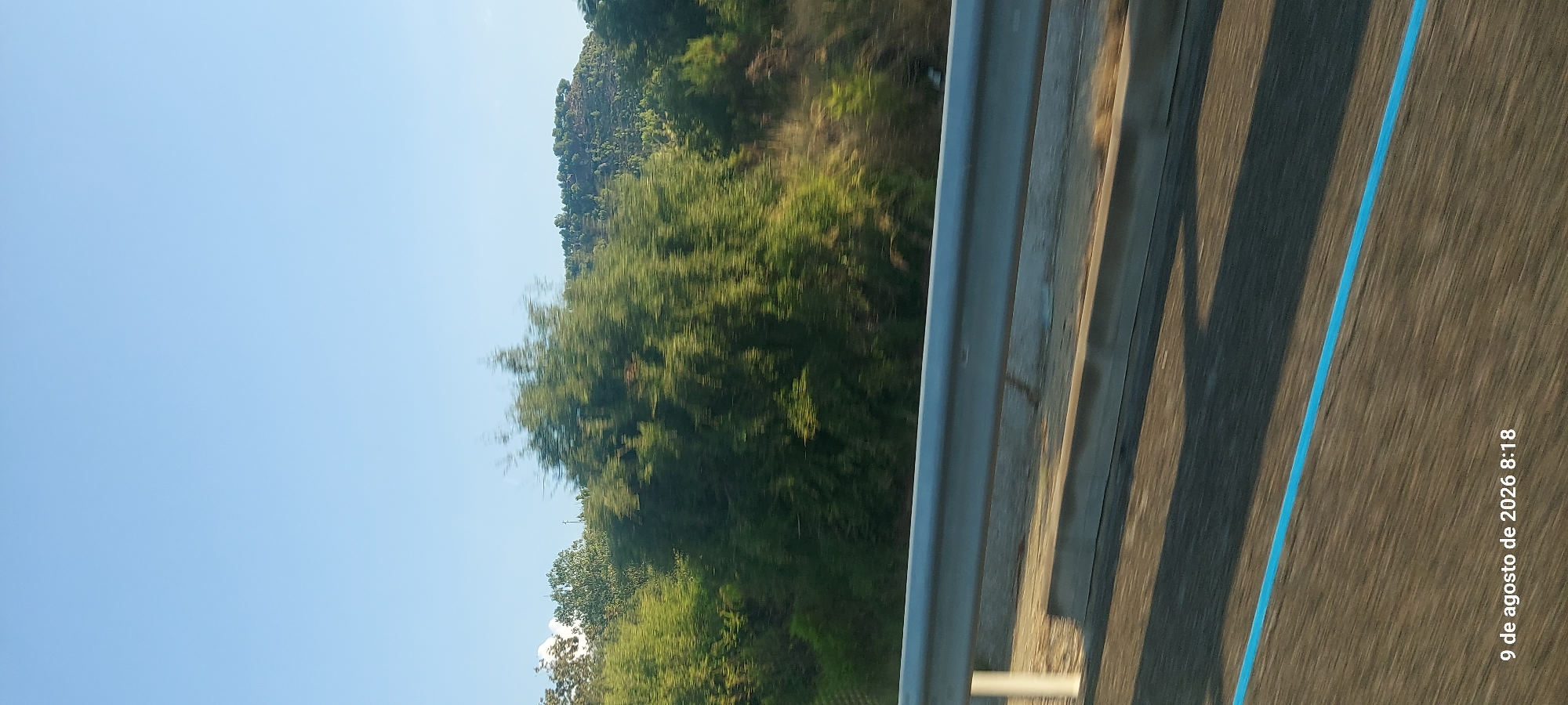 The image shows a view from a moving vehicle on a road, likely a highway, in l'Espluga de Francolí, Spain. The foreground is dominated by the road surface, which has a distinct blue line running across it. A metallic guardrail is visible above the road. In the middle ground, there are blurred green trees and vegetation, indicating motion. The background features a gently sloping hill covered in more trees and sparse vegetation, under a clear blue sky. The timestamp in the lower left corner reads "9 de agosto de 2026 8:18," which translates to "August 9, 2026 8:18." No people or specific landmarks of l'Espluga de Francolí are identifiable due to the motion blur and the nature of the landscape.