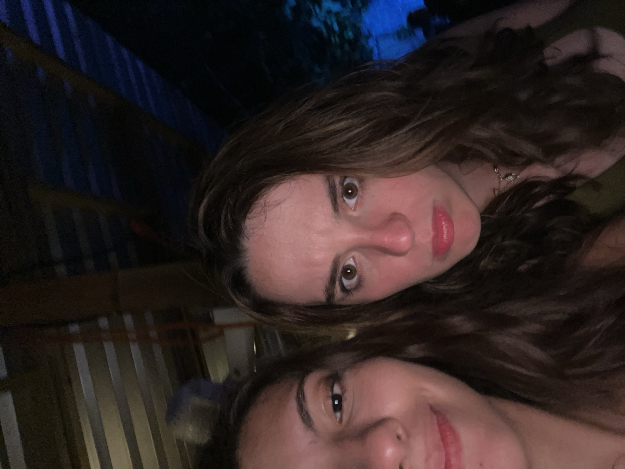 Two women are close together in an indoor setting, appearing to take a selfie. The woman on top has light skin, brown hair, and brown eyes; she is wearing lipstick. The woman on the bottom has tan skin, brown hair pulled back, and brown eyes; she also wears lipstick. The location is likely Puerto Escondido, Mexico, although specific details to confirm this within the image are not present. The background is dark, with indistinct structures and a blue light source visible in the upper portion. The foreground is also dark.