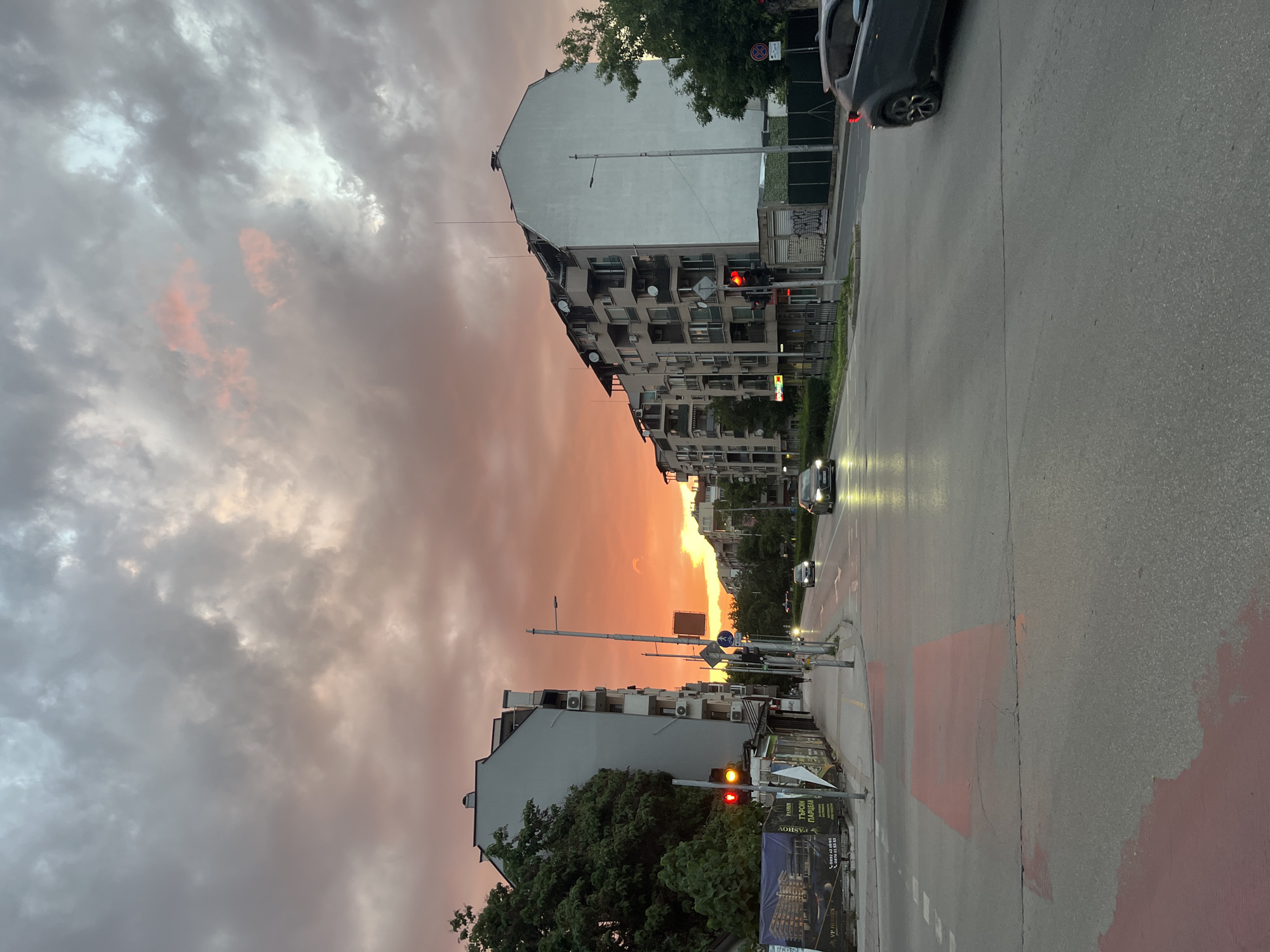 The image shows a street scene in an urban setting. The sky is filled with clouds and tinged with orange and pink hues, indicating either a sunrise or sunset. Buildings line both sides of the street, with some appearing to be apartment blocks featuring multiple windows and balconies. Streetlights are visible, and traffic lights are set to red. Cars are present on the road, with some moving. The road surface is asphalt and includes markings such as painted sections. A poster or advertisement is positioned at the roadside. Trees are scattered throughout the scene, adding greenery to the urban environment.