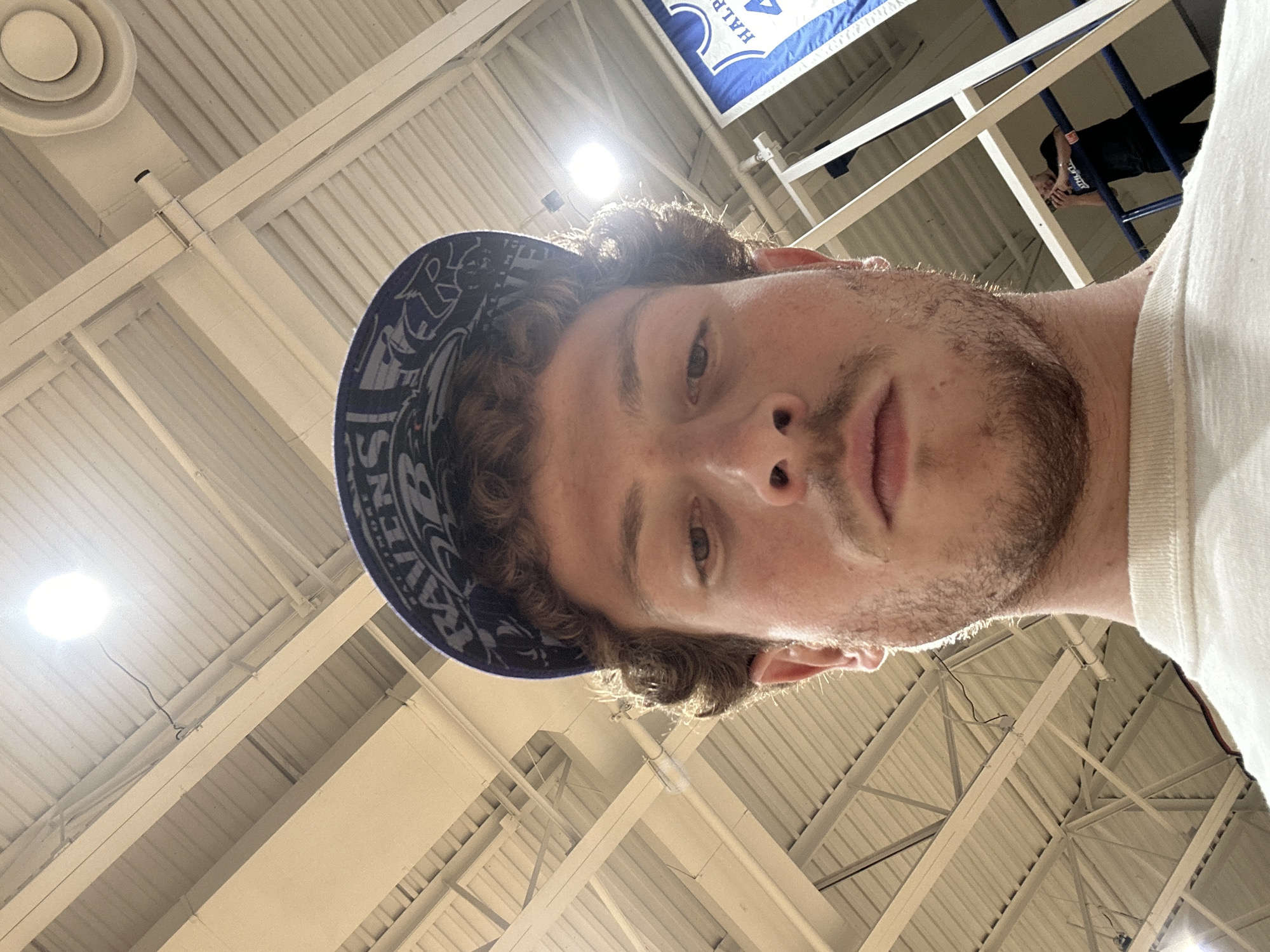 An indoor, low-angle selfie captures a young man looking upwards, occupying the majority of the frame, with the building's ceiling structure and another person in the background. The image is rotated 90 degrees clockwise.
The main subject, a man with light brown, curly hair and stubble/short beard, wears a dark blue cap with a white pattern and a white t-shirt. He gazes directly into the camera with a neutral expression. Bright artificial lights from the ceiling create a strong backlight, outlining his hair.
The setting appears to be a large public indoor facility, possibly a gymnasium or sports arena, characterized by high ceilings with exposed white metal beams and rafters. A circular air vent is visible in the top left. Another bright, circular light fixture is prominent on the left, casting a strong glare.
In the upper right background, a second individual is seen standing on an elevated platform or bleachers, wearing a dark top. Their features are not distinct.
A blue and white banner hangs from the ceiling structure, and the word "HALE" is partially visible in white lettering on the blue section. Wires or cables are also seen running along the ceiling.
The activity is a selfie being taken. The overall impression is one of being inside a well-lit, functional building. The location context indicates this is in New York, United States of America.