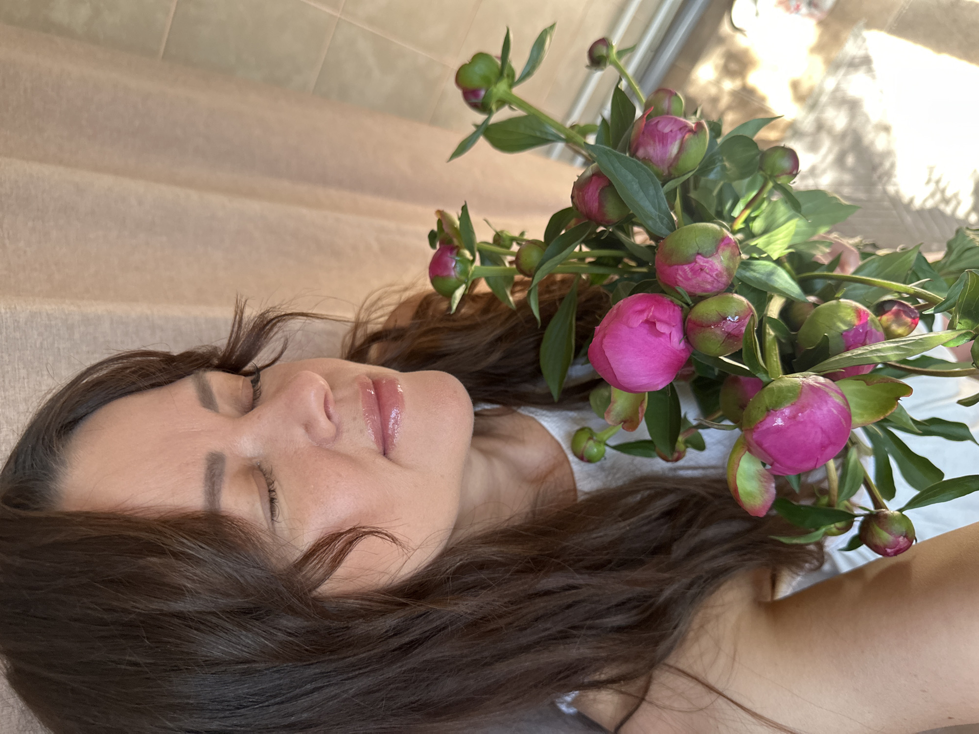A medium shot photograph shows a woman with dark brown hair and closed eyes, holding a bouquet of pink peonies. She is wearing a white top. The background consists of a light-colored curtain and a window with sunlight streaming through, casting shadows on the floor. The image is taken indoors, possibly in a residential setting in Chaikovsky, Russia, given the natural lighting and domestic environment.