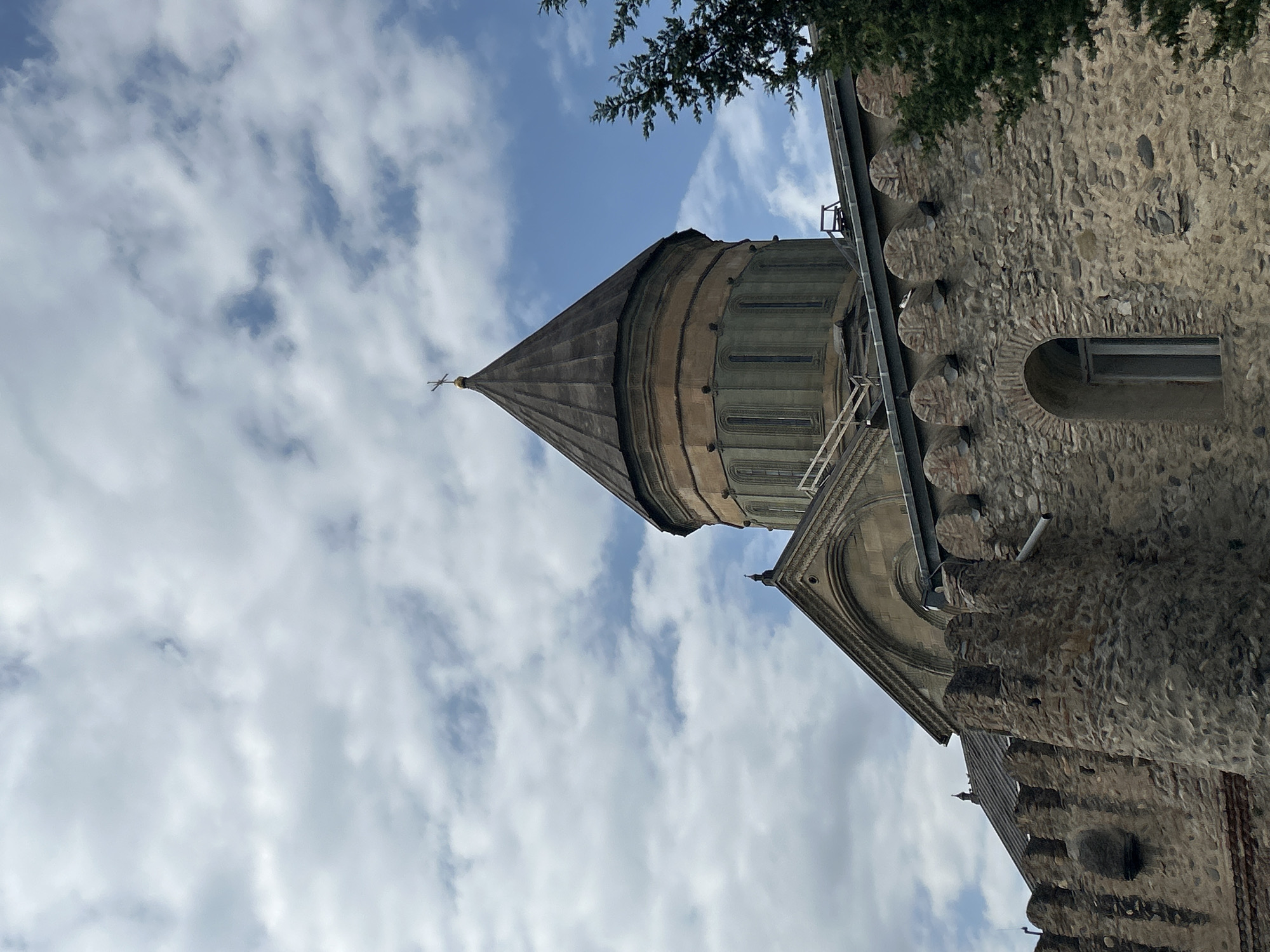 The image shows the Svetitskhoveli Cathedral in Mtskheta, Georgia. The cathedral is made of stone and has a large dome with a cross on top. The sky is cloudy with patches of blue visible. The foreground shows a stone wall with an arched window. There are some trees on the right side of the image. The scene is calm and serene, with no people visible. The overall impression is one of historical significance and architectural beauty.