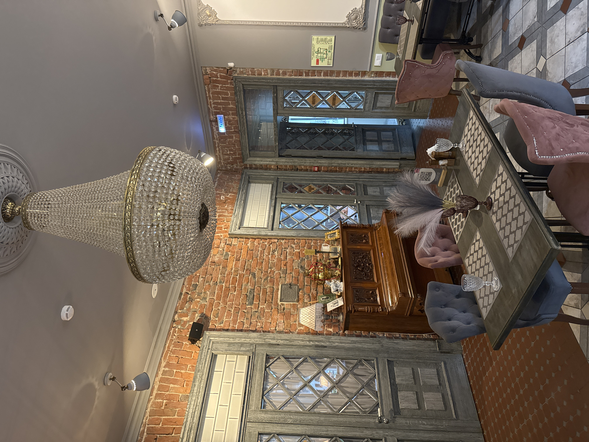 The image shows a dimly lit dining area with an ornate chandelier hanging from the ceiling. The walls are a mix of exposed red brick and grey plaster. Several wooden doors with diamond-patterned windows are visible. A long wooden table is set with glasses, napkins, and a decorative feather arrangement in a vase. Around the table, several upholstered chairs in muted tones of pink and grey are arranged. To the left of the table, a wooden cabinet with intricate carvings stands against the brick wall. In the background, a sign with Russian text is visible on the wall, and a fire exit sign with Cyrillic text is illuminated by a blue light. The overall atmosphere appears to be that of a restaurant or cafe.