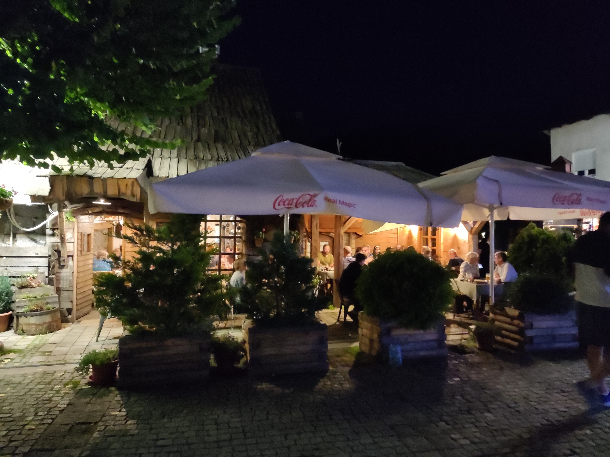 The image shows an outdoor seating area of a restaurant or cafe at night. Several patrons are seated at tables under umbrellas. One umbrella has a Coca-Cola logo and the text "Real Magic." The building behind the seating area appears to be made of wood with a thatched or shingled roof. There are also several potted plants and trees around the seating area. The ground is paved with cobblestones. The scene suggests a cozy and relaxed atmosphere, likely in Kolašin, Montenegro, given the context provided.