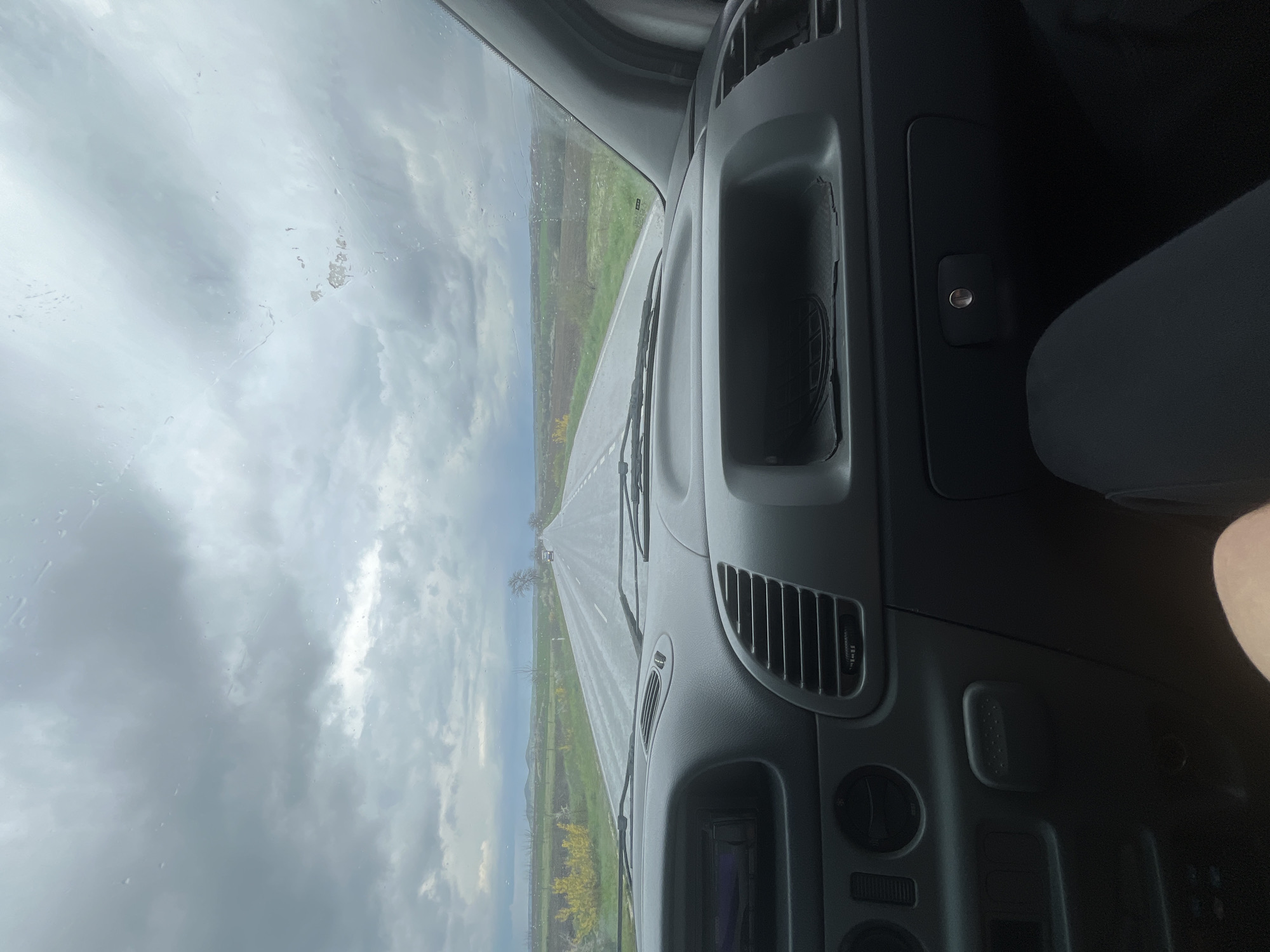 A view from inside a vehicle driving on a road towards Byala Reka, Bulgaria. The gray dashboard of the vehicle is prominent in the foreground. The road ahead is straight, flanked by greenery and fields. The sky is overcast with dark, heavy clouds. The windshield is wet, possibly from rain. A passenger's leg is partially visible on the right side of the dashboard.