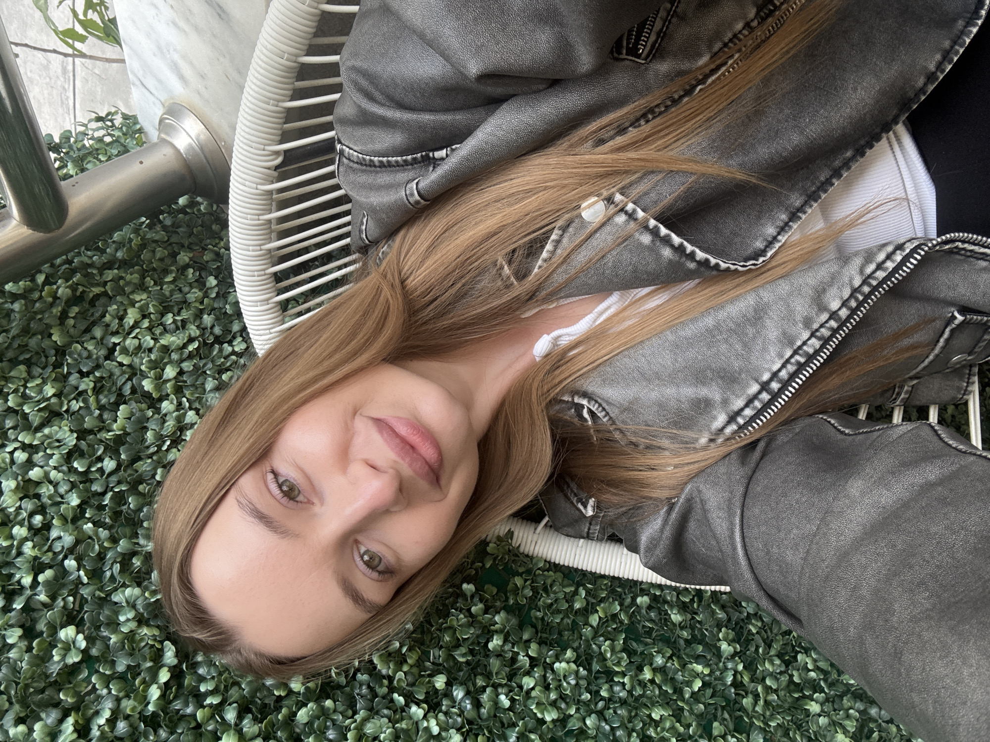 A fair-skinned woman with light brown hair poses for a photo while seated in a white wicker chair. She is wearing a gray leather jacket over a white top. The chair is placed on a green artificial foliage surface, which could be a wall covering or ground cover. To the left, a metallic object, possibly a chair leg or fixture, is visible. The setting implies an outdoor cafe or lounge, but specific city location details are absent. The photograph has been taken from an overhead angle.