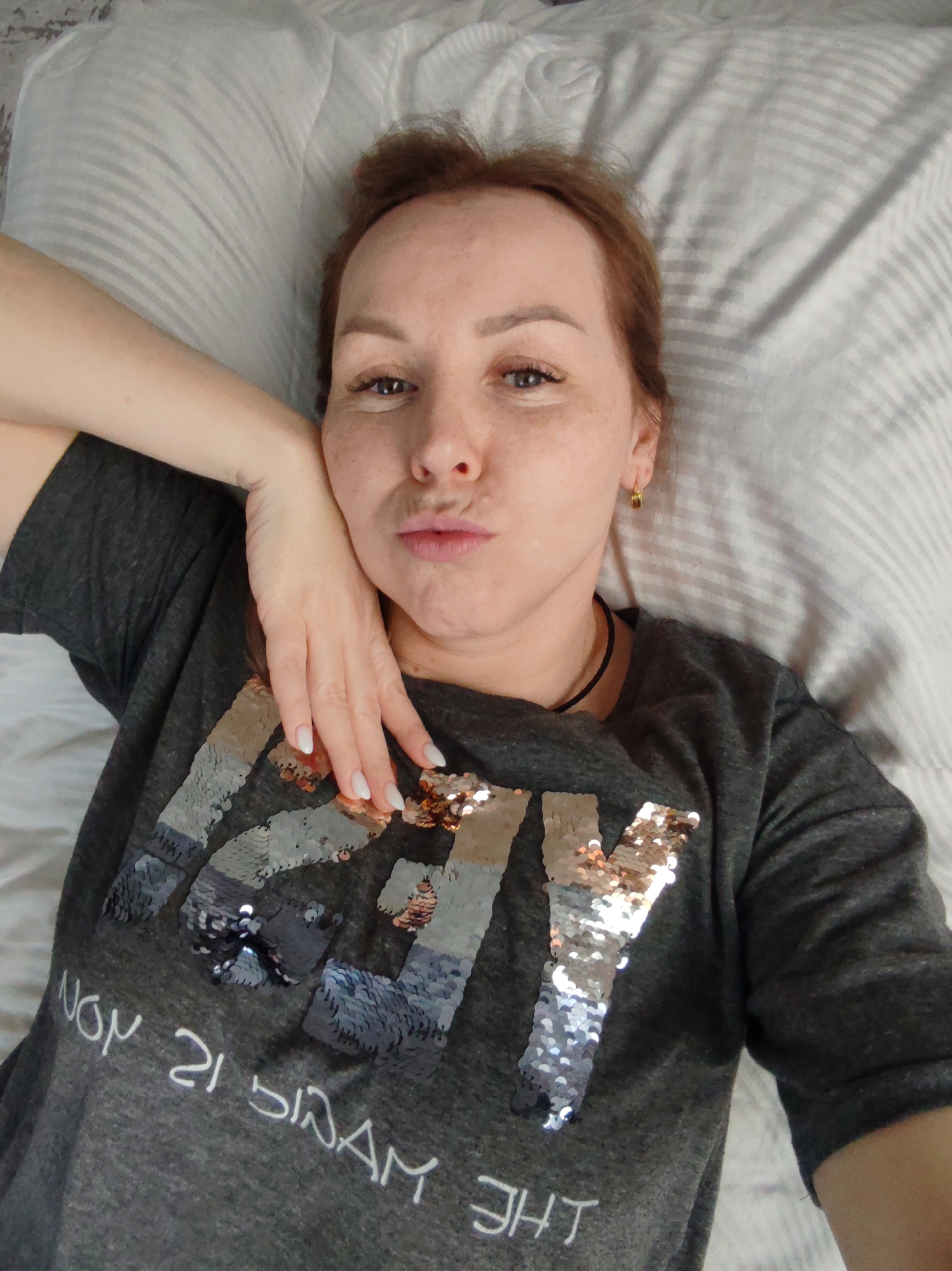 A woman lies on a bed or couch, facing upwards towards the camera, taking a selfie. She has reddish-brown hair pulled back, and fair skin with freckles visible across her face. Her left hand is raised to rest against her cheek and jawline. She is looking directly at the camera with a playful, pouting expression, commonly known as a "duck face."
She is wearing a dark gray short-sleeved t-shirt adorned with a large, sequined design on the front. The sequins are reversible, appearing in silver and rose gold/bronze tones, forming large, blocky letters. Below the sequined letters, white text is printed, which appears reversed in the image, likely due to a front-facing camera or mirroring. When read normally, this text says "THE MAGIC IS ON." She is also wearing small, gold-colored hoop earrings and a thin black choker necklace. Her nails are manicured with a light, possibly white, polish.
The background consists of a light-colored pillow, likely white or light gray, with subtle horizontal stripes. The overall setting appears to be indoors, suggesting a relaxed environment like a bedroom. There are no specific cues within the image to determine the time of day or external location, although the context indicates Chaikovsky, Russia.
