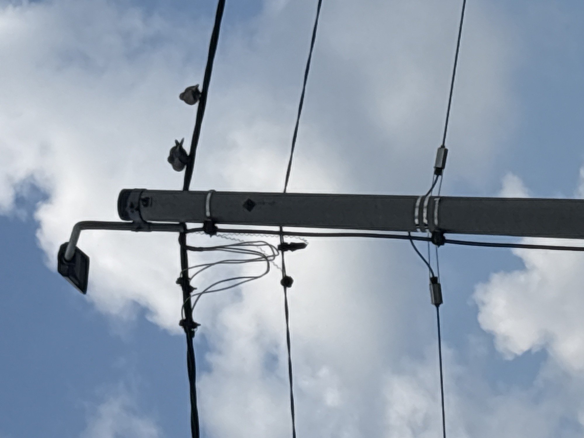 The image shows a utility pole in the city of Drożki, Poland. The pole is tall and gray, with electrical wires attached to it at various heights. Three birds are perched on one of the wires. A street lamp is mounted on top of the pole. The sky is blue with some white clouds. The scene is typical of an urban or suburban environment.