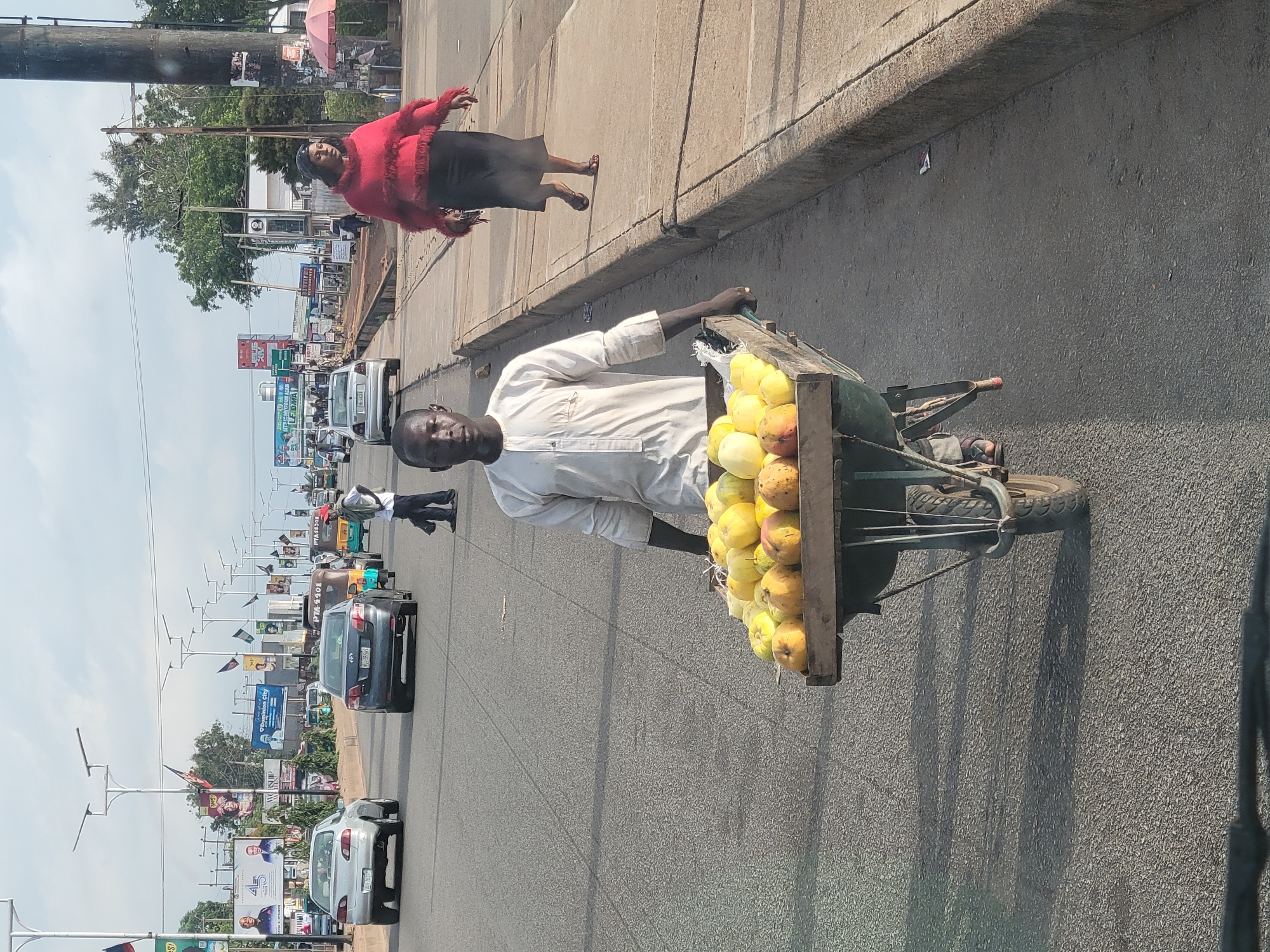 The image depicts a street scene in Jos, Nigeria. A man in a light-colored shirt is pushing a wheelbarrow filled with yellow fruits, likely mangoes or similar produce. He is walking on the road, with traffic visible in the background. A woman in a red top and dark skirt is walking on the sidewalk. The street is lined with billboards and streetlights, indicating a commercial area. Cars and other vehicles are moving along the road. The sky is overcast.