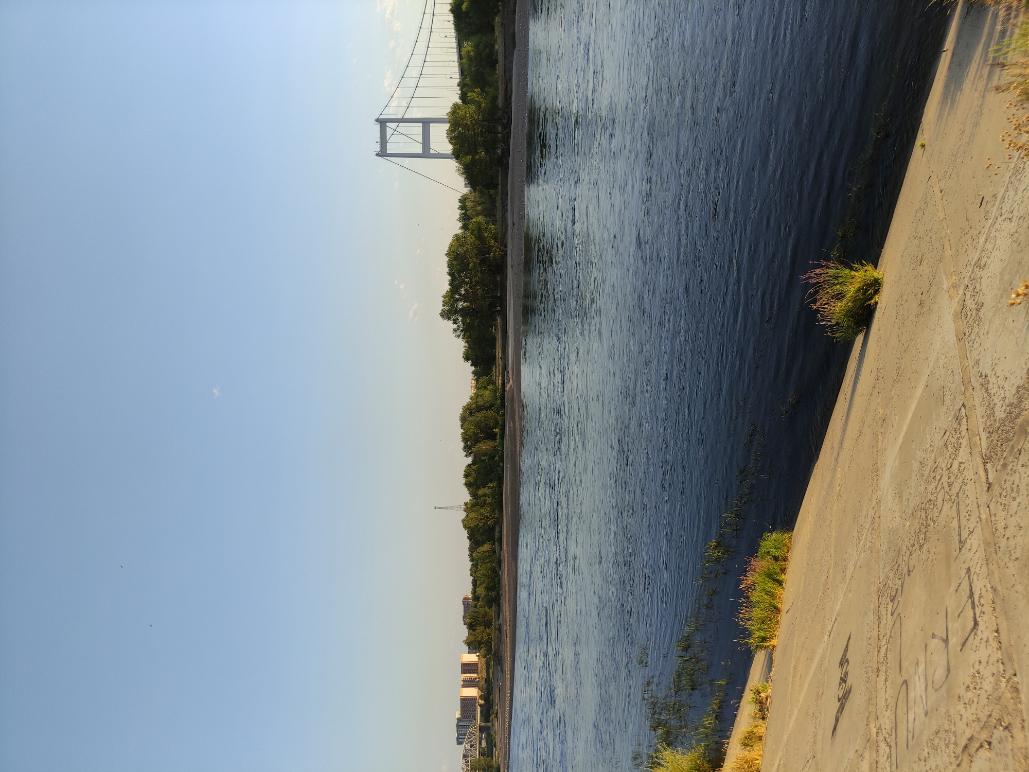 The image depicts a wide, calm river with a sandy bank on the left side. The bank is lined with concrete and some vegetation. Across the river, there is a line of trees, and beyond that, several buildings and a radio tower are visible. A large suspension bridge spans the river in the distance on the right side of the image, and another bridge is partially visible on the left. The sky is clear blue with a few small clouds and two birds flying. The scene appears to be set in the city of Semey, Kazakhstan, likely near the Irtysh River, given the presence of a suspension bridge and the general landscape. The lighting suggests it is late afternoon or early evening, with the sun casting long shadows. There are no people visible in the image.