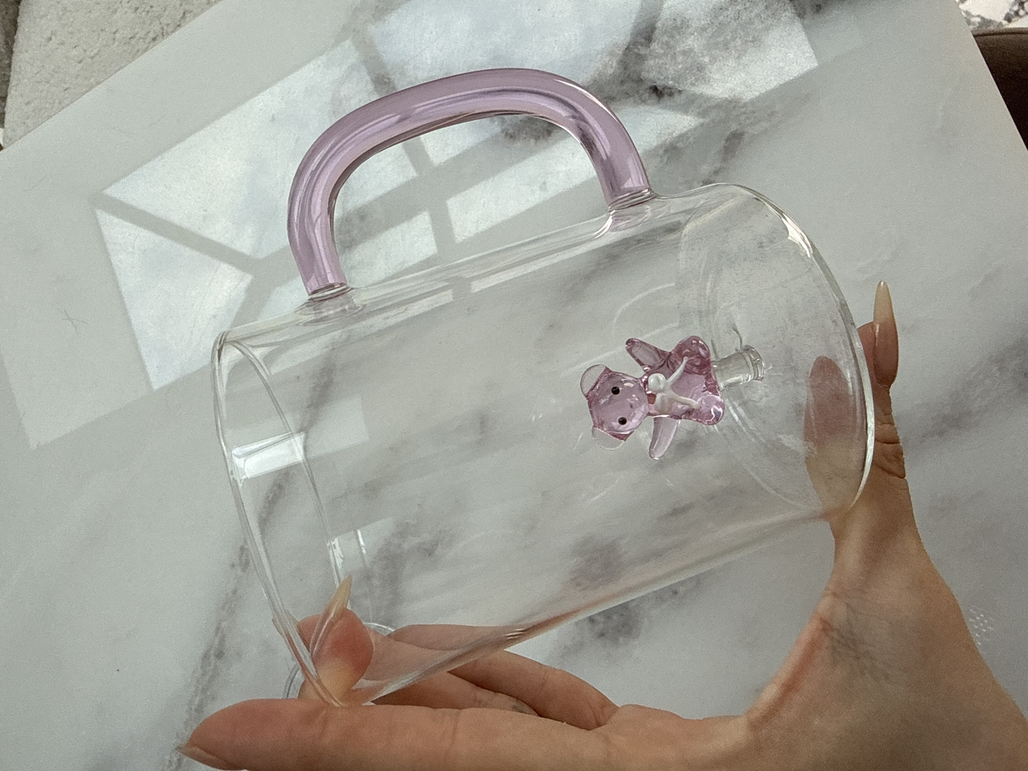 A human hand holds a clear glass mug with a pink handle and a pink glass bear inside against a background of white marble. The location is not discernible from the image and no information suggests Nottingham, United Kingdom.