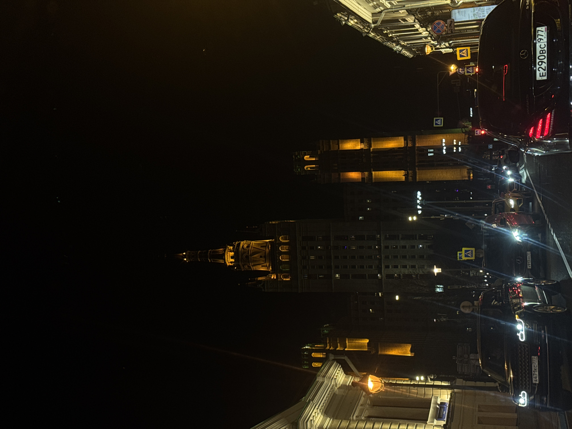 The image captures a vibrant night scene of a city street in Moscow, showcasing architectural landmarks and urban activity. The time of day is clearly night, characterized by the dark sky and the dominance of artificial lighting.
On the left side of the frame, towering over the scene, is a prominent illuminated building with distinctive ornate architecture, featuring a central spire and classical detailing. This structure strongly resembles one of Moscow's famous "Seven Sisters" Stalinist skyscrapers, such as the Hotel Ukraina or the building on Kudrinskaya Square. Adjacent to it are other buildings, also illuminated with warm light from within their windows, suggesting ongoing activity.
The right side of the image features a bustling street with several cars. In the immediate foreground, a black Mercedes-Benz Maybach is clearly visible, facing away from the viewer. Its red taillights are illuminated, and the Russian license plate "E290BC 977 RUS" is clearly readable, with "977" indicating a Moscow registration. The word "MAYBACH" is also visible on the car's trunk. Further down the street, a line of cars with bright headlights and red taillights suggests ongoing traffic. Another car in the mid-ground has a partially visible license plate "A471MC 777 RUS".
Street furniture includes numerous traffic signs mounted on poles, such as a "No Stopping" sign (red circle with diagonal line) and "Pedestrian Crossing" signs (blue square with white triangle and person). Streetlights with a warm, orange glow illuminate the pavement and parts of the buildings. No specific people are clearly discernible, but the presence of many cars implies a populated urban environment. The weather appears clear, with no rain or fog. The overall impression is one of an active, well-lit metropolis at night.