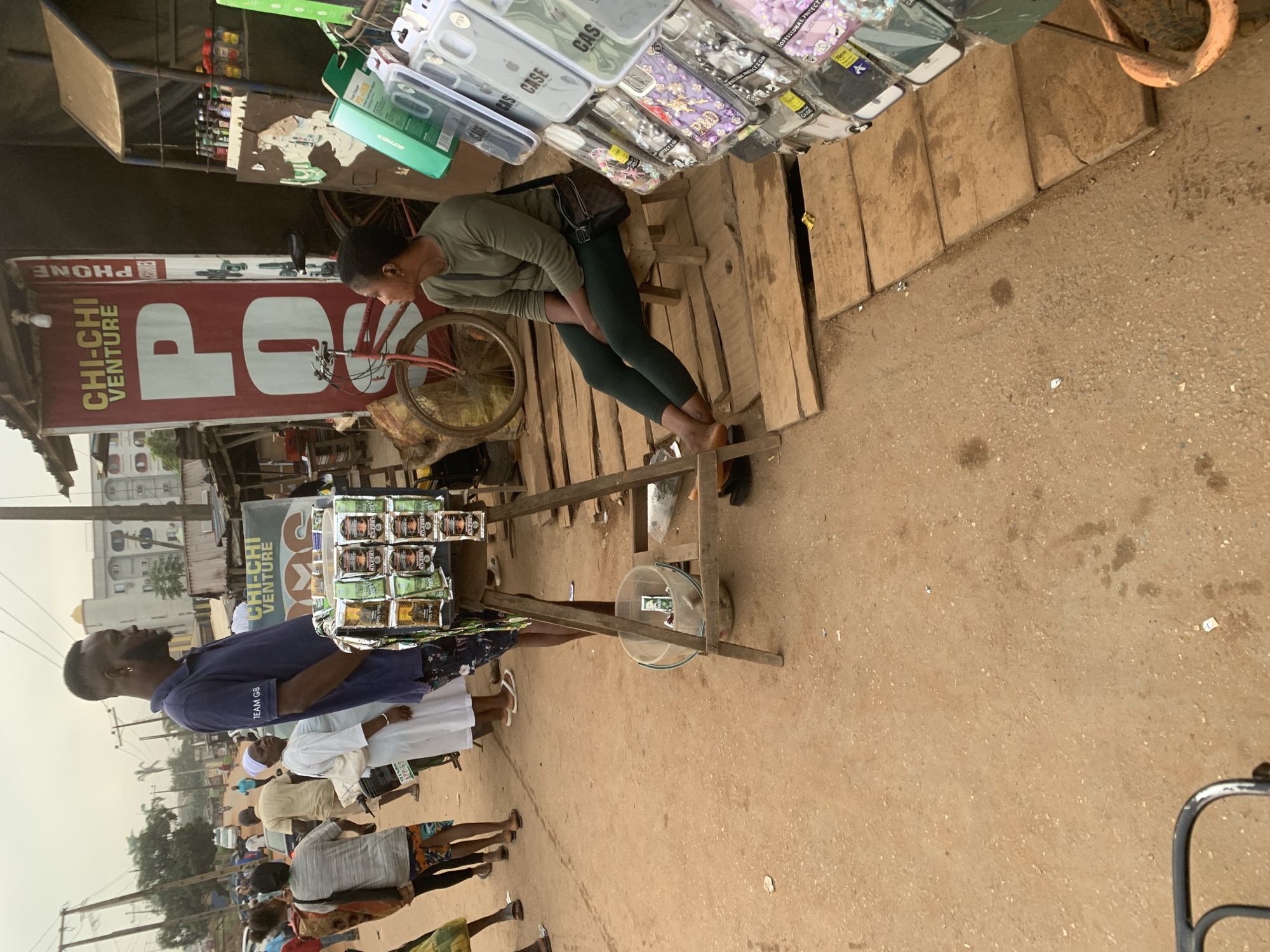In Shagamu, Nigeria, a market scene unfolds. A tall man with a fade haircut, wearing a dark blue polo shirt with "TEAM G8" printed on it and patterned shorts, stands next to a display of small, bagged items, possibly snacks or toiletries. He appears to be a vendor. In the background, a woman wearing a green long-sleeved shirt and dark leggings sits on a low wooden stool, facing away from the camera. She has a crossbody bag with a checkered pattern. Beside her is a red bicycle. In front of the man is a clear plastic bucket with some items inside. The marketplace is characterized by a dirt ground, wooden planks forming a walkway, and makeshift stalls. A large red banner with "CHI-CHI VENTURE" and "PHONE" printed in white and yellow dominates the background, suggesting it is a stall selling mobile phone accessories. Other stalls display phone cases and various bottled products. Pedestrians are visible in the distance, with a building in the far background. The sky is overcast.