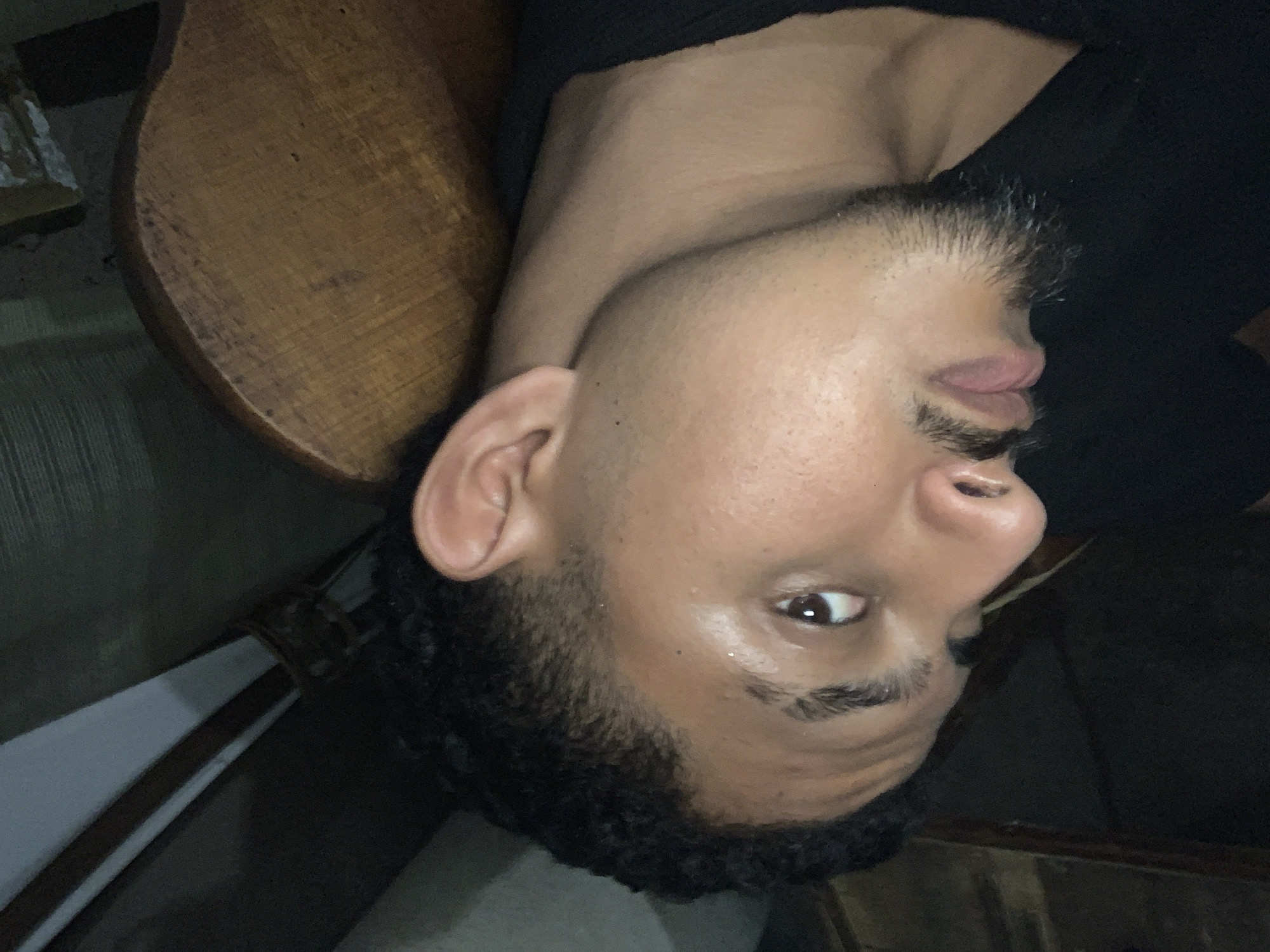 The image shows a close-up, head-and-shoulders shot of a person with dark, curly hair and a goatee. The person is wearing a black shirt and is looking slightly upwards and to the left, with their head tilted. The background is dimly lit and appears to be indoors, with elements of what looks like furniture or wooden structures visible. The lighting is low, with highlights on the person's skin, particularly on the forehead and cheek. There is a hint of a wooden chair or bench behind the person. The specific location in Santa Ana, Venezuela, cannot be determined from the visual information alone.