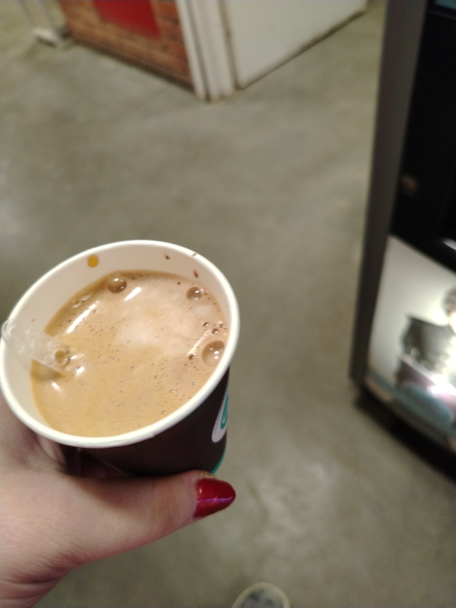 The image captures a close-up, slightly blurred shot of a hand holding a paper cup filled with a frothy, light brown beverage, likely coffee or hot chocolate. The hand, with fair skin, displays red or maroon nail polish on one finger. A clear plastic stir stick is visible inside the cup. The cup itself is white with a dark brown or black base, and a small portion of a light blue or teal design or logo is visible on its side.
The setting appears to be indoors. The background is out of focus, showing a light gray, possibly concrete or linoleum, floor. In the upper left, a blurry section of a reddish-brown brick wall with white trim or a doorframe is visible. To the right, a dark, metallic structure resembling part of a vending machine or an industrial appliance stands vertically. The overall lighting is dim and uniform, suggesting indoor conditions without direct natural light. No specific activity beyond holding the drink is depicted, and no emotions are discernible. There is no visible text due to the blurriness and angle.