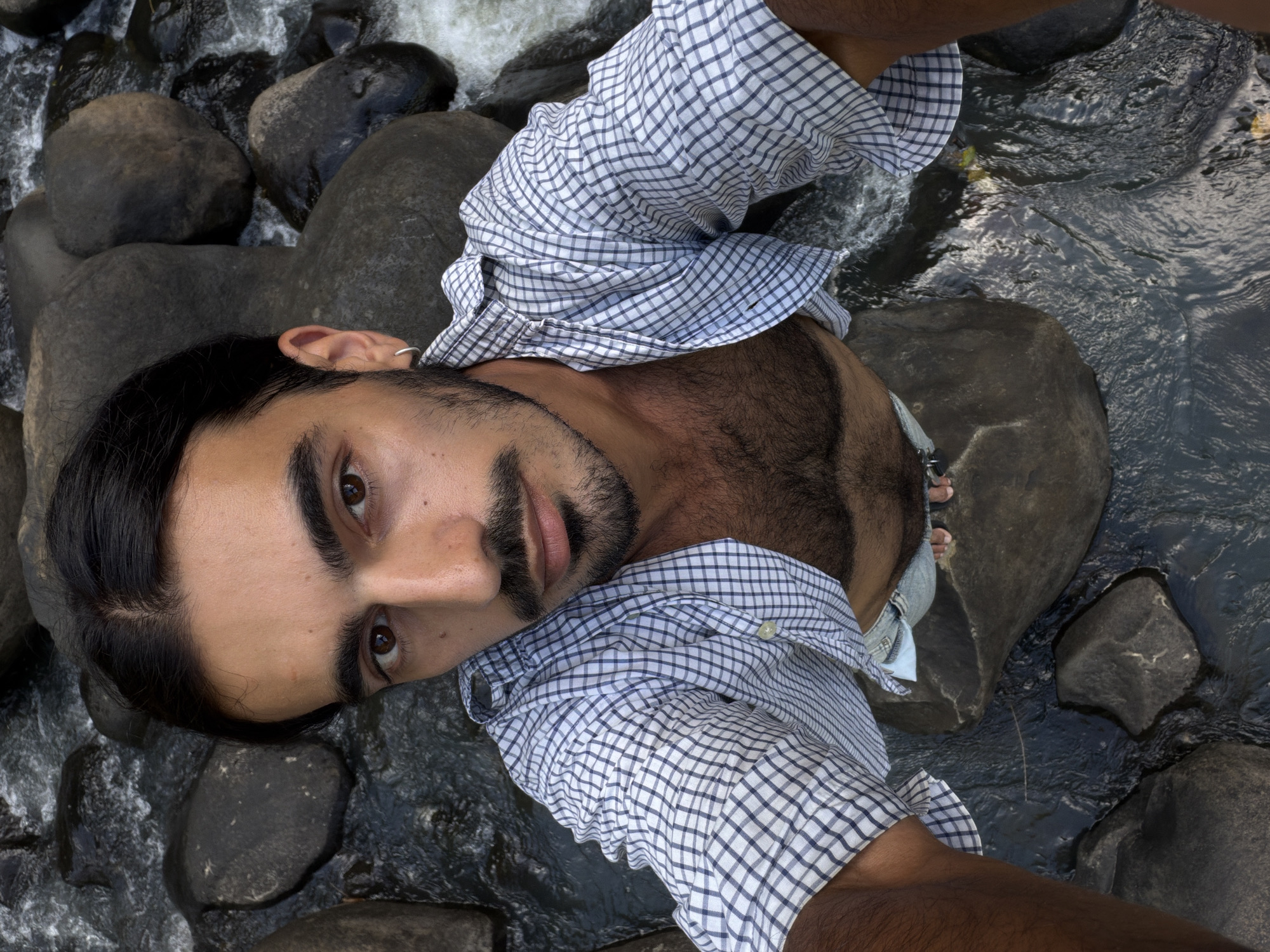 A man is lying on rocks in a riverbed. The riverbed contains smooth, dark stones and flowing water. The man is wearing a blue and white checkered shirt, unbuttoned to reveal his chest. His feet, clad in denim, are partially submerged. He has dark hair, a beard, and is looking directly at the camera. The location is Selemadeg Timur, Indonesia.