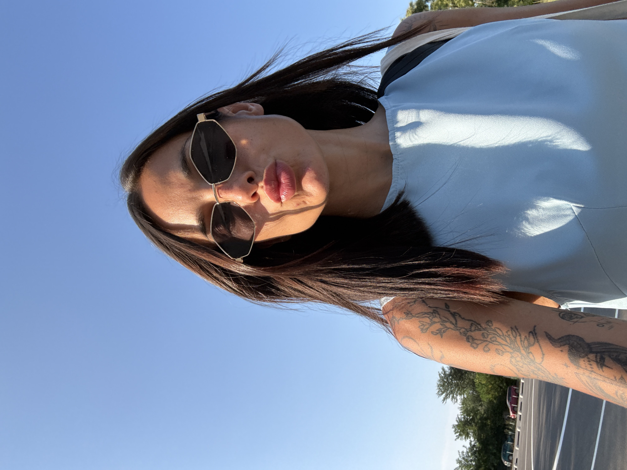 In the city of Pleven, Bulgaria, a woman is captured in a close-up selfie against a clear blue sky. She is wearing large, angular sunglasses and a light blue top. Her lips are pursed in a kiss gesture, and her long, dark hair is slightly windswept. Visible on her left arm are several intricate tattoos, including a floral design and what appears to be a mythological figure. The background shows a parking lot with a few cars and a line of trees, suggesting an outdoor, possibly urban or suburban setting. The lighting indicates it is daytime, with the sun casting shadows on her face and clothing.