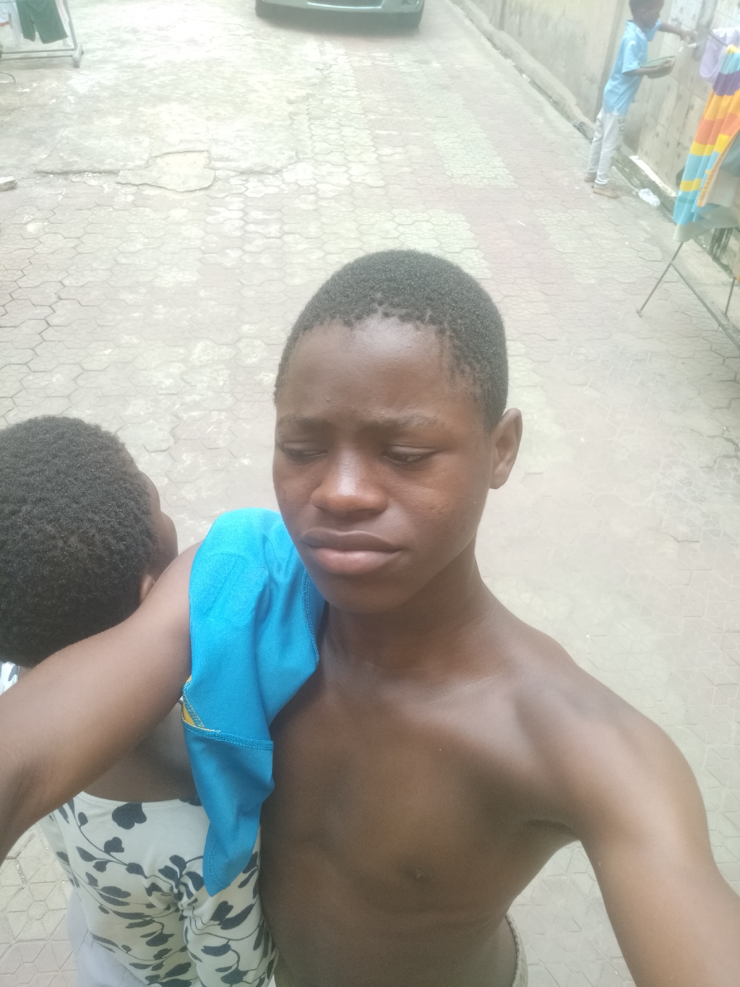 A close-up outdoor photograph features a young Black person (likely male, a child or pre-teen) in the foreground, looking downwards with a serious expression. Their head and upper torso are visible, and they are shirtless, with a bright blue cloth draped over their left shoulder. Their short, dark hair is visible. Another young Black child, wearing a white top with a dark leaf pattern, is partially visible behind the main subject, with only the back of their head and shoulder visible.
The background shows a paved ground made of hexagonal interlocking blocks, which appears somewhat worn and cracked in places. To the right, further back, another young person, also appearing to be a child or pre-teen, is standing next to a wall. This individual is wearing a light blue long-sleeved shirt and light-colored trousers, and appears to be engaged in an activity near the wall, possibly washing something or interacting with a water source. A colorful clothes drying rack, holding various items, is positioned to the right of this person. In the far background, a part of a dark-colored car is visible, and the scene suggests an outdoor, possibly residential, setting like an alleyway or courtyard. The lighting is bright, indicating daytime. There is no visible text in the image. The scene conveys everyday life in Shangisha, Nigeria.