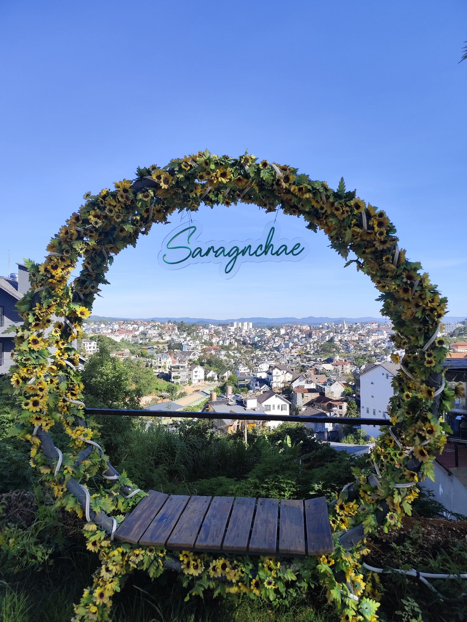 The photograph features a circular archway adorned with sunflowers, framing a panoramic view of a densely populated cityscape under a clear blue sky. In the center of the arch, a translucent sign with the word "Saragnchae" in cursive script is suspended. The arch itself is constructed from a dark, cylindrical material and decorated with a dense arrangement of artificial sunflowers, interspersed with green leaves and white ribbon-like accents. The base of the arch incorporates a wooden bench.
The background reveals a sprawling city composed of closely packed buildings, primarily houses and low-rise structures, with occasional taller buildings visible in the distance. The urban landscape is interspersed with patches of green vegetation, creating a contrast between the built environment and natural elements.
Given the "Saragnchae" sign and architectural style of the houses, it is strongly suggestive that the image was taken in Da Lat, Vietnam.