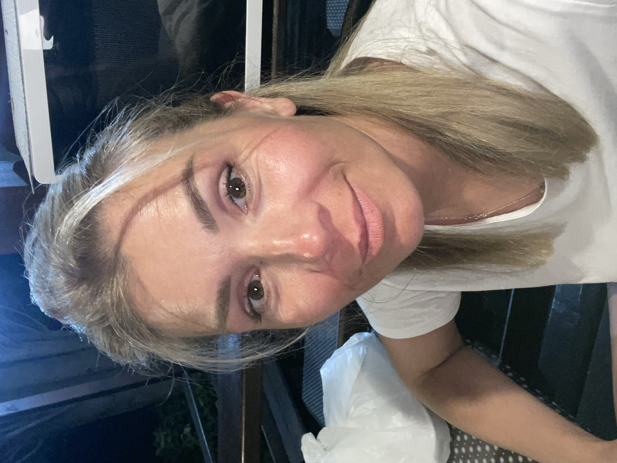 The original image is a close-up, selfie-style photograph of a blonde woman with light skin and brown eyes. She is wearing a white t-shirt and has light makeup on her eyes and lips. The woman is looking directly at the camera and smiling slightly. Her hair is pulled back loosely, with some strands falling around her face. In the background, there is a white plastic bag on a dark, textured surface to her left. To her right, there appears to be a dark, outdoor setting with some wooden structures and a white object that might be part of an appliance. The lighting suggests it is evening or night, with artificial light sources illuminating the woman. There are no visible geographical markers to indicate a specific city.
