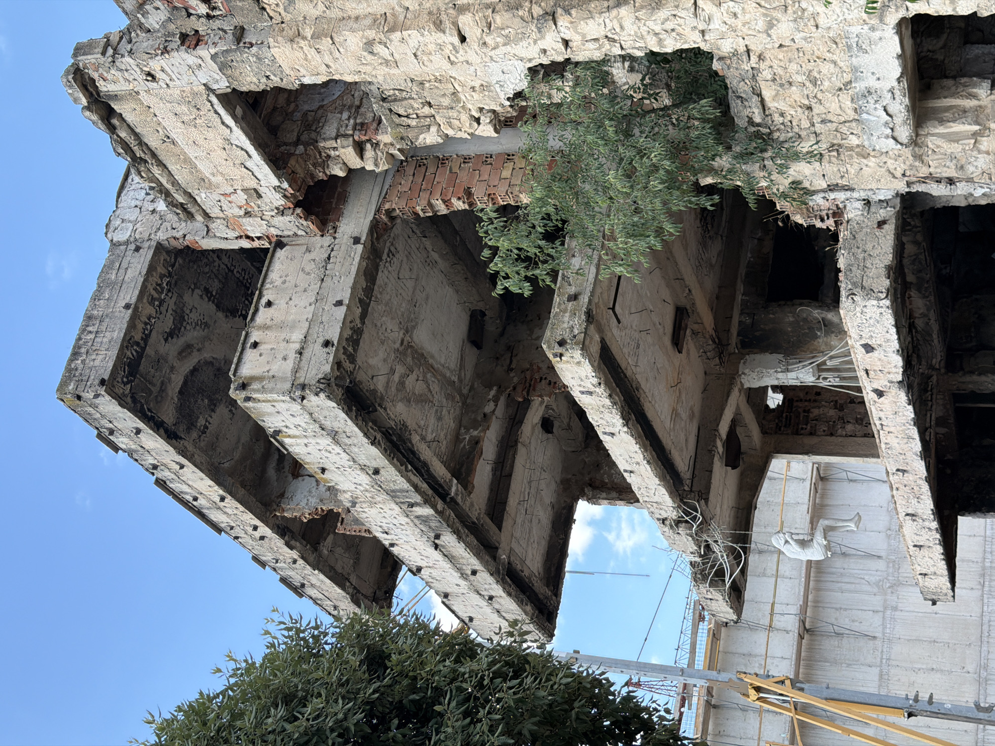The image shows the partially destroyed facade of a building in Mostar, Bosnia and Herzegovina. The building is made of concrete and stone, with several stories visible. The concrete floors are broken and crumbling, revealing exposed rebar. The stone walls are cracked and weathered. Greenery is growing out of the damaged structure, with trees and bushes visible on several levels. In the foreground, on the left side of the image, a large tree with green leaves is present. On the right side, the stone facade of the building is more prominent, showing significant damage and wear.
In the mid-ground, a white, human-like sculpture is hanging from wires, appearing to be seated on an unseen object and possibly swinging. This sculpture is positioned on a concrete slab that juts out from the building. Behind the sculpture and to the left, scaffolding and construction materials are visible, suggesting ongoing work or demolition. The sky is clear and blue with a few faint clouds. The overall scene depicts a state of ruin and decay, juxtaposed with elements of nature reclaiming the structure and signs of potential rebuilding or artistic intervention.