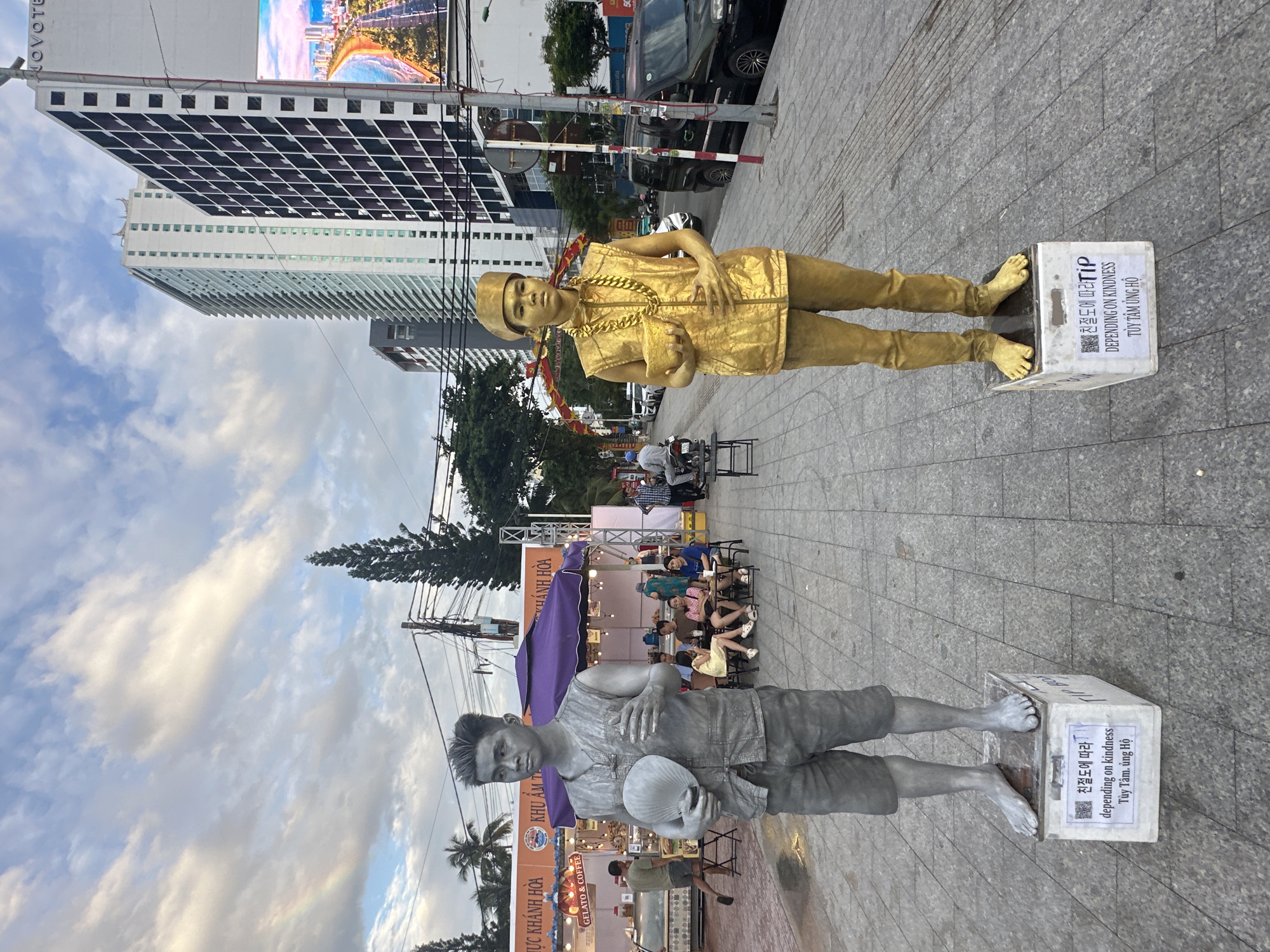 An upright, eye-level street photograph captures two "living statues" performing in an urban setting, likely in Nha Trang, Vietnam, as suggested by the context.
In the foreground, a gold-painted street performer stands on a white box, wearing a sleeveless top, pants, and a cap, adorned with a large gold chain and holding what appears to be a small pouch. To their left, a silver-painted performer, similarly poised on a white box, wears a sleeveless top and shorts, holding a small hat or bowl. Both performers maintain stoic expressions.
The white boxes serve as tip containers, bearing identical text in Korean ("친절도에 따라"), English ("DEPENDING ON KINDNESS"), and Vietnamese ("TÙY TÂM ỦNG HỘ" - roughly "Support as you wish" or "Depending on your heart's desire"), along with the word "TIP" and a QR code.
The background reveals a bustling street scene with several people, some seated at outdoor tables, suggesting cafes or food stalls. Visible signs include "GELATO & COFFEE" and "KHU ẨM THỰC KHÁNH HÒA" (Khánh Hòa Food Area). Cars and motorcycles are parked or moving on the street. Towering buildings, including one prominently displaying "NOVOTEL" at its top, define the cityscape. Overhead, numerous power lines crisscross the sky. The sky is partly cloudy, indicating daytime, possibly late afternoon. The overall atmosphere is lively, characteristic of a tourist or commercial district.