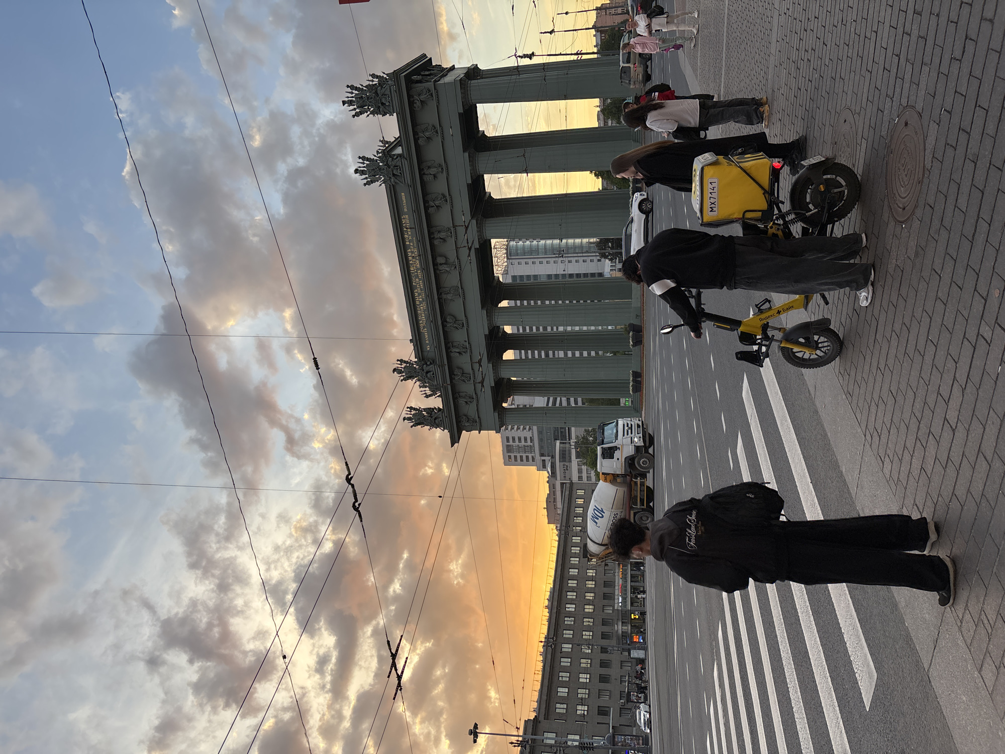 The image captures a vibrant late afternoon or early evening street scene in Saint Petersburg, Russia, centered around the historic Narva Triumphal Arch. The sky is dramatic, filled with grey and white clouds that glow with warm golden and orange hues from the setting sun, contrasting with patches of pale blue sky. Numerous overhead power lines crisscross the upper part of the frame.
In the foreground and mid-ground, several people are present on a paved sidewalk and a zebra crossing. A person in a black jacket and pants stands on the right, facing away from the viewer. Closer to the center, another individual in dark clothing stands next to a bright yellow electric scooter. The scooter is equipped with a large yellow delivery bag on its rear, bearing the text "Я.ЕДА" (Yandex.Food) and a license plate "MX7141". The scooter's frame also shows the branding "Яндекс Банк" (Yandex Bank). Other pedestrians are visible further back on the sidewalk.
The prominent architectural subject is the Narva Triumphal Arch, a large classical structure with imposing columns, intricate sculptural details, and statues atop its pediment. A long inscription in Russian is visible on the arch, which reads: "ПОБЕДОНОСНОМУ РОССИЙСКОМУ ВОЙСКУ И ХРАБРОМУ НАРОДУ СООБЩИВШЕМУ МИР И СПОКОЙСТВИЕ ЕВРОПЕ" (To the victorious Russian army and brave people who brought peace and tranquility to Europe). Behind and through the arch, modern city buildings, including one with many windows, and a white truck are visible. The scene conveys a blend of historical grandeur and contemporary urban life under a beautiful sunset sky.