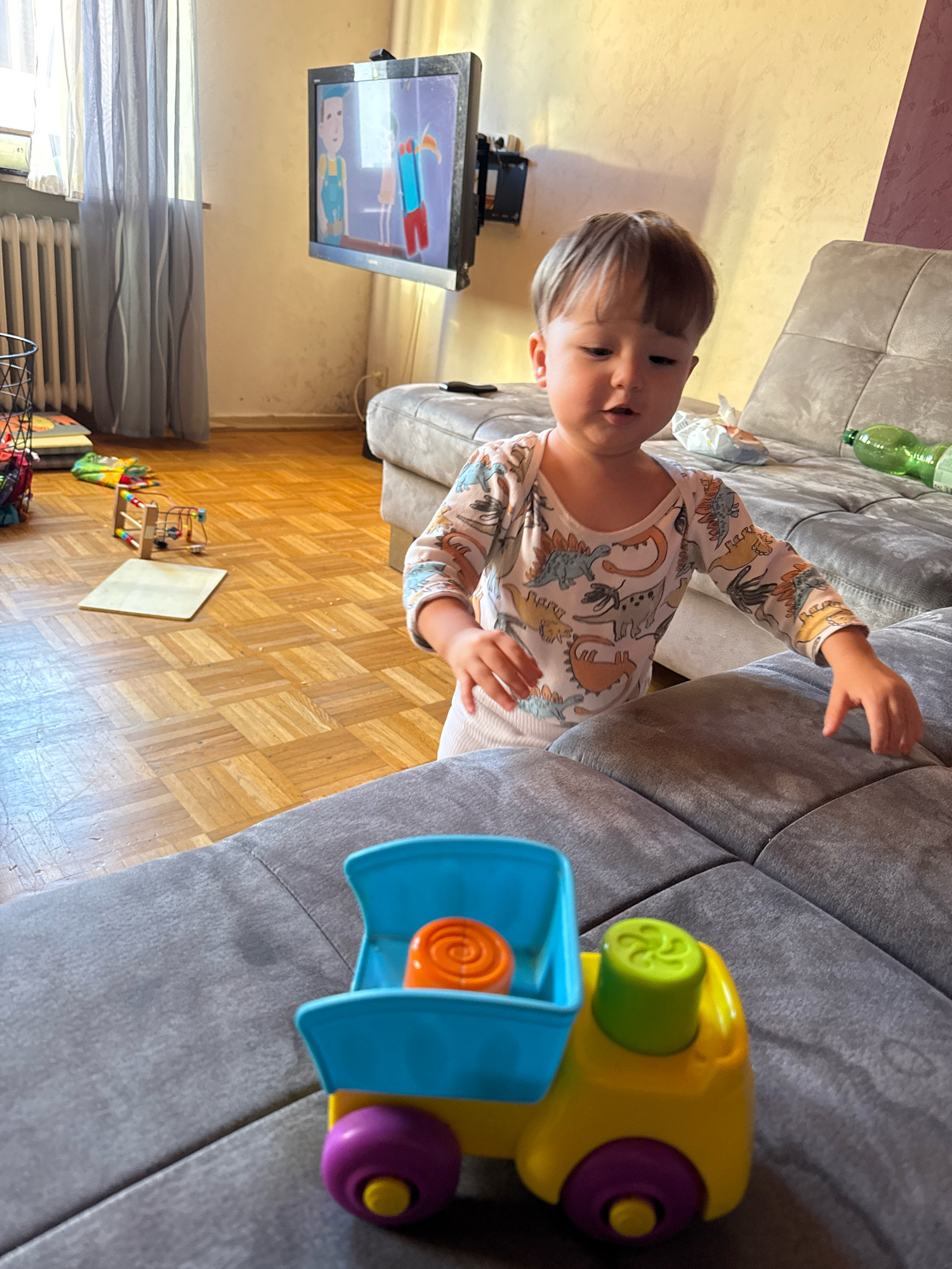 The image depicts a child's playroom or living area, possibly located in Überherrn, Germany, as suggested by the prompt. In the foreground, a toddler wearing a long-sleeved white onesie adorned with a dinosaur pattern is standing and looking downwards. The child's hands are visible, and they appear to be interacting with a toy truck that rests on a grey sofa. The toy truck is colorful, featuring a blue bed, a yellow cab, and purple wheels. Inside the truck bed, an orange object with a spiral pattern is visible.
The background reveals a grey sectional sofa, part of which the child is leaning on. Further back, a television screen mounted on the wall displays a cartoon with two figures. To the left, a window is covered by a grey sheer curtain, and a radiator is visible beneath it. The floor is made of polished wood with a parquet pattern. In the middle ground, on the floor, various children's toys are scattered, including a wooden bead maze toy and a few stacked items. A green plastic bottle is also present on the sofa. The overall lighting suggests daytime, with natural light filtering in from the window.