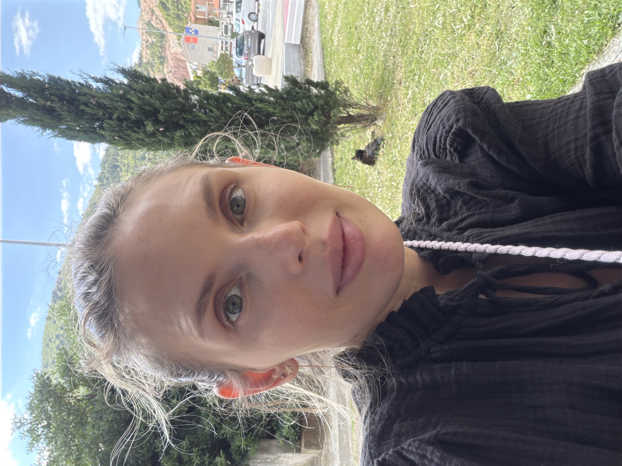 A woman is taking a selfie outdoors. She has fair skin, blonde hair, and light-colored eyes. She is wearing a black top with a white drawstring. In the background, there is greenery, a tree, and buildings. The street scene includes cars and street signs. It is Budva, Montenegro due to the Montenegrin flag visible in the background.