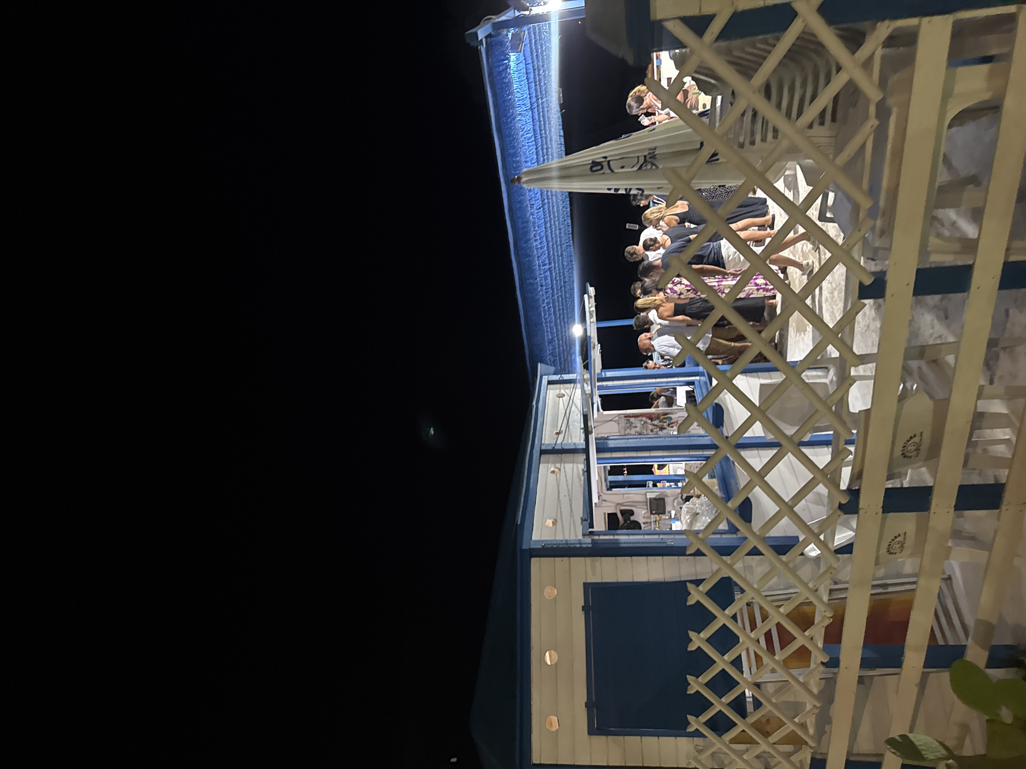 This is a nighttime photograph taken outdoors at a beach bar in Isola delle Femmine, Italy. The scene is lit by artificial lights. In the foreground, a white lattice fence with blue accents runs horizontally across the frame. Behind the fence, a small, blue and white building with round windows and a blue shuttered door is visible. To the right of the building is an open area where a group of people are gathered. They appear to be standing around a counter or bar. A large white umbrella with a cartoonish face is propped up in the middle of the gathering. The background is a dark night sky. The ground appears to be sandy or paved. The overall atmosphere suggests a casual social gathering at a beachside establishment.