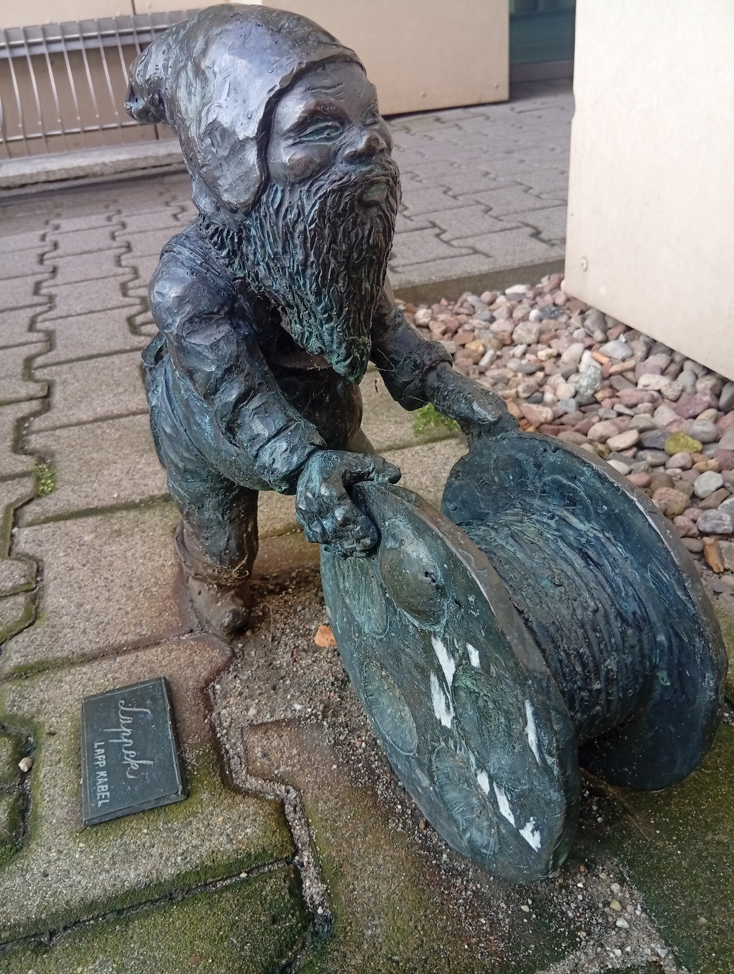 A bronze statue of a dwarf is positioned on a paved walkway in Wroclaw, Poland. The dwarf is depicted in a squatting posture, holding two large, circular objects that resemble spools or coils. It has a long beard and wears a hat, characteristic of the many dwarf statues found throughout the city. In front of the statue, a small, rectangular plaque is embedded in the pavement. The plaque has text that reads "Lappek" in script and "LAPP KABEL" below it. The walkway is made of interlocking concrete pavers, some of which are covered with moss. The area surrounding the walkway is covered with small stones. In the background, a building with a light-colored facade is visible, along with a metal railing.