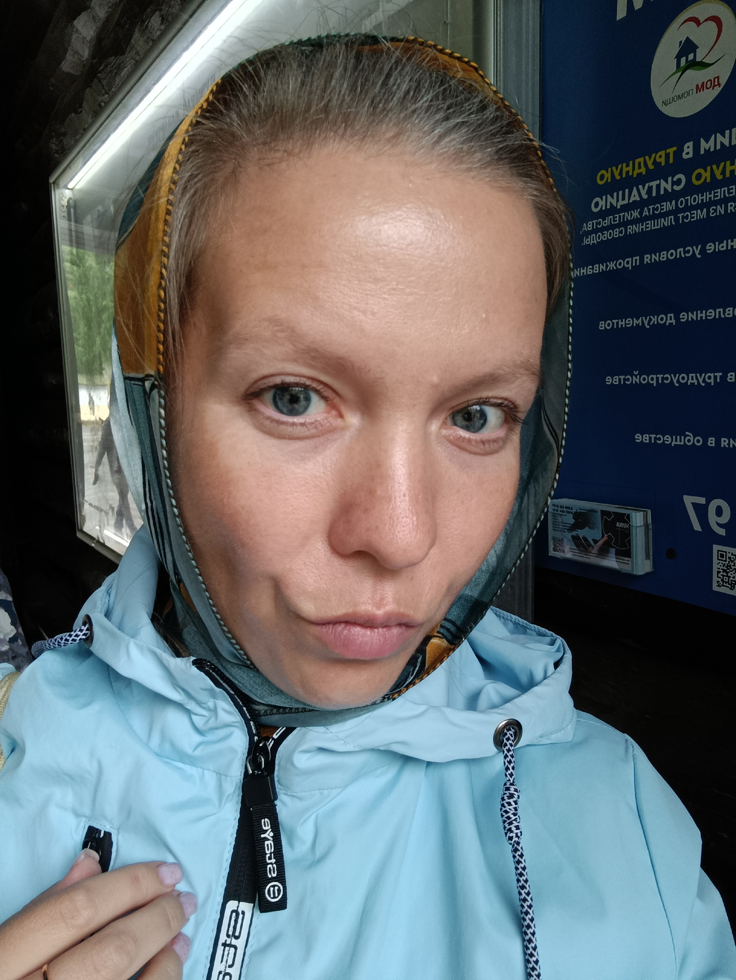 A young woman with light skin, blue eyes, and graying blonde hair pulled back is captured in a close-up, frontal portrait. She is wearing a light blue hooded jacket with a black zipper and a patterned scarf that covers her hair. Her lips are pursed. The background features a dark, textured surface on the left and a blue panel with text in an unfamiliar script on the right, along with a circular logo depicting a house and heart. The overall setting appears to be outdoors, possibly near a bus stop or public transportation area, given the informational signage. No specific landmarks of Yekaterinburg are visible.