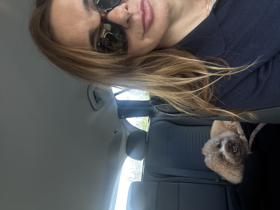 The image displays the interior of a private vehicle, with the specific location within Alcobaça, Portugal, not discernible from the visual evidence. The foreground is dominated by the partial view of an adult female, positioned as if capturing a self-portrait. She has light brown hair, wears dark tortoiseshell-framed sunglasses, and a dark blue collared top. A gold chain is visible on her neck. Her lips are slightly parted, revealing a light pink color. In the background, a small, light brown, curly-haired dog is resting on a dark textile covering the rear passenger seat. The dog's mouth is open, exposing its teeth. The vehicle's interior features dark-colored rear seats with headrests and a light grey headliner. Daylight is visible through the rear windows, suggesting an outdoor environment beyond the vehicle.