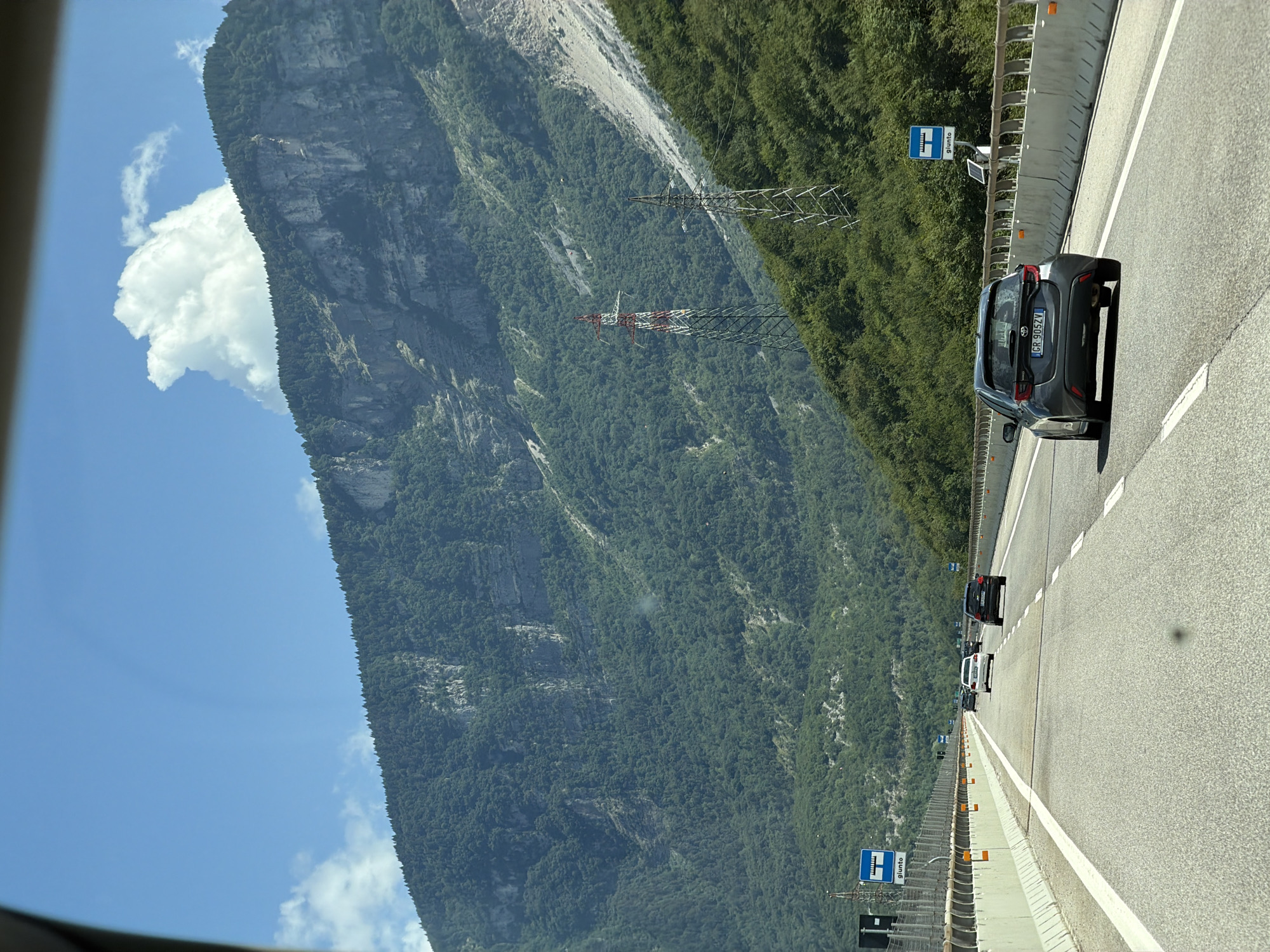 A grey 2020 Toyota Yaris with license plate GR 905ZV is driving on a multi-lane highway that cuts through a mountainous, forested area. The highway has concrete barriers and metal guardrails. In the background, a large, steep mountain covered in dense green trees rises towards a clear blue sky with a few white clouds. There are also two large electrical transmission towers visible on the mountainside. Road signs are visible on the left side of the highway, and other cars are driving ahead. The perspective suggests the photo was taken from a vehicle moving forward on the highway.