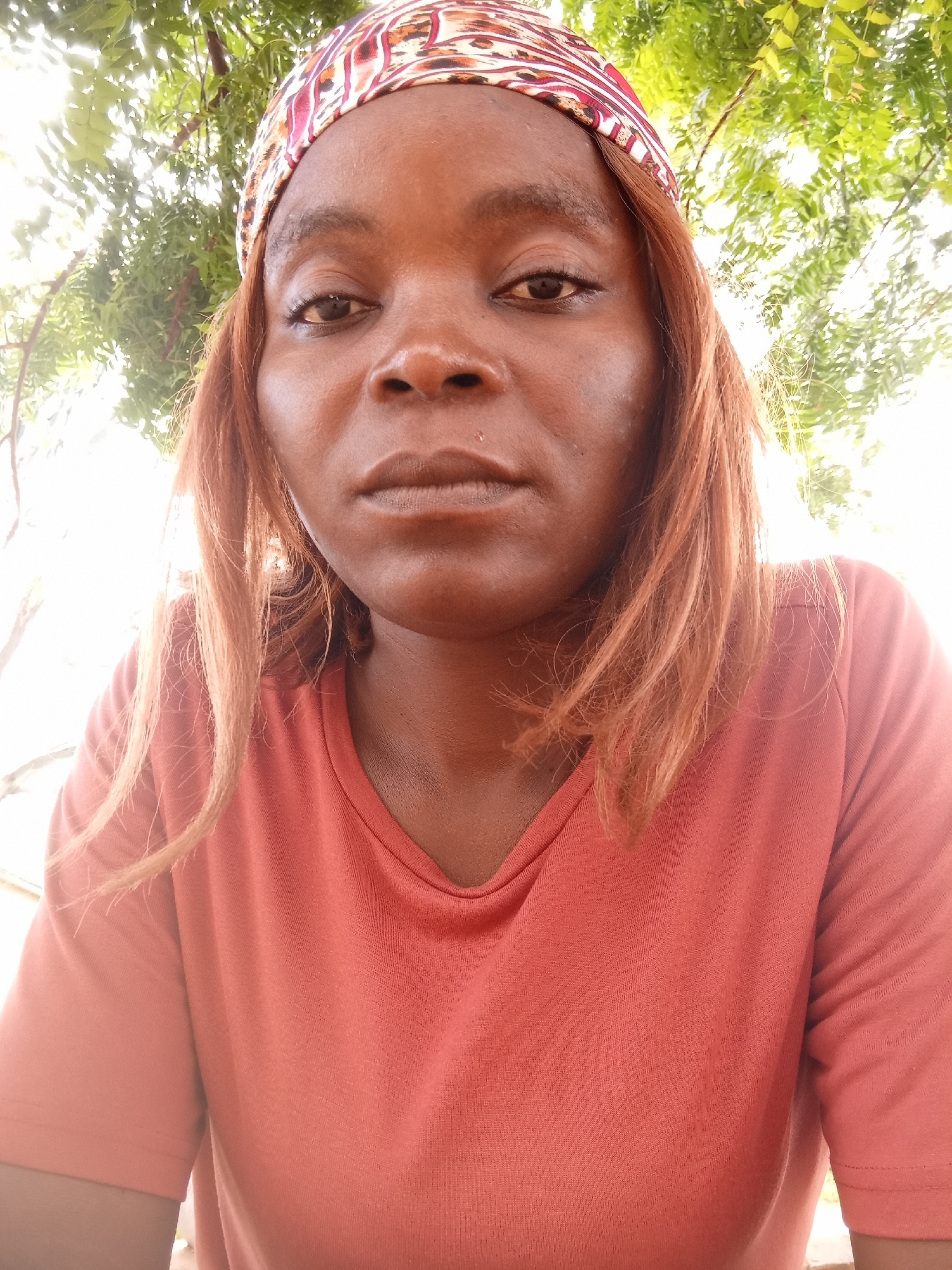 A medium-shot, chest-up, outdoor selfie of a Black woman wearing a coral-colored t-shirt and a headwrap. She has light brown hair and is looking directly at the camera with a neutral expression. The background consists of green foliage and bright, hazy sunlight. The image appears to have been taken in Kason, Nigeria.